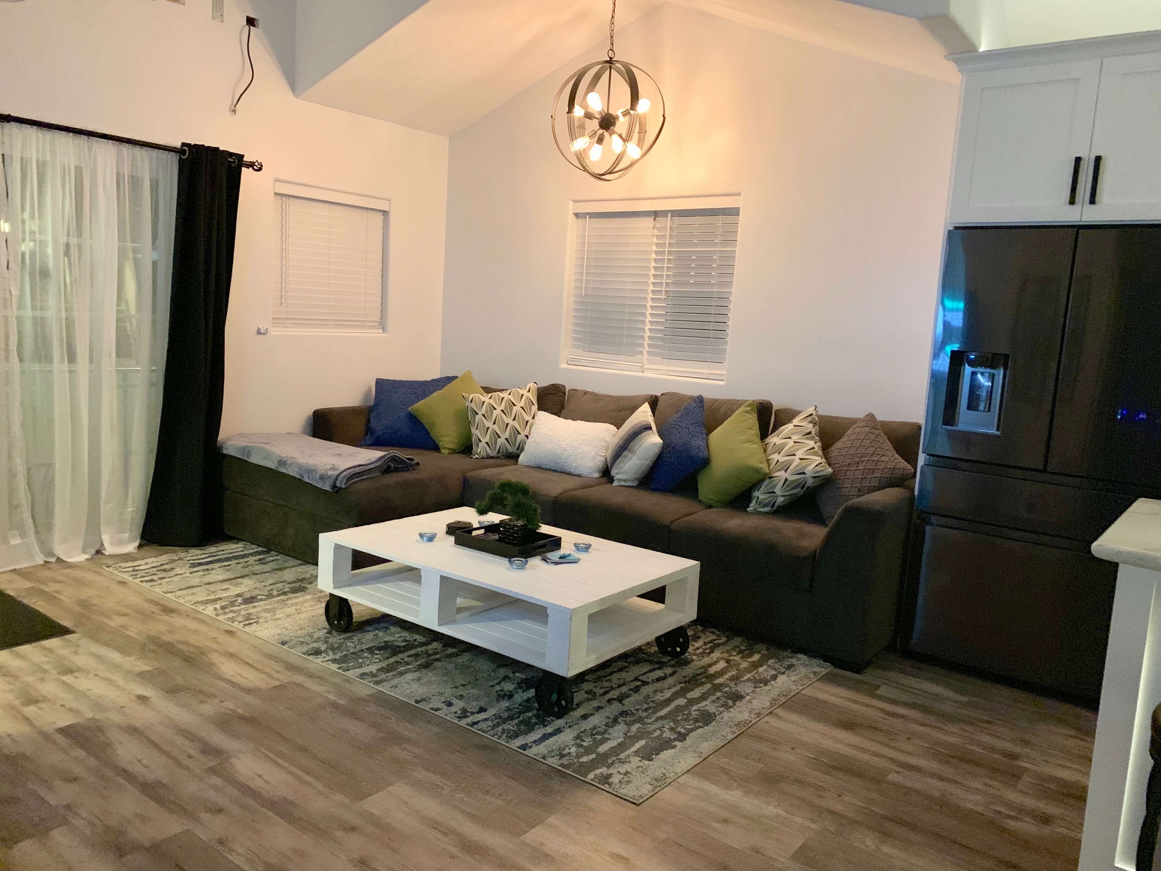 The image shows a living room with a large sectional sofa covered in various colored pillows, a white coffee table on wheels, and a modern light fixture hanging from the ceiling.