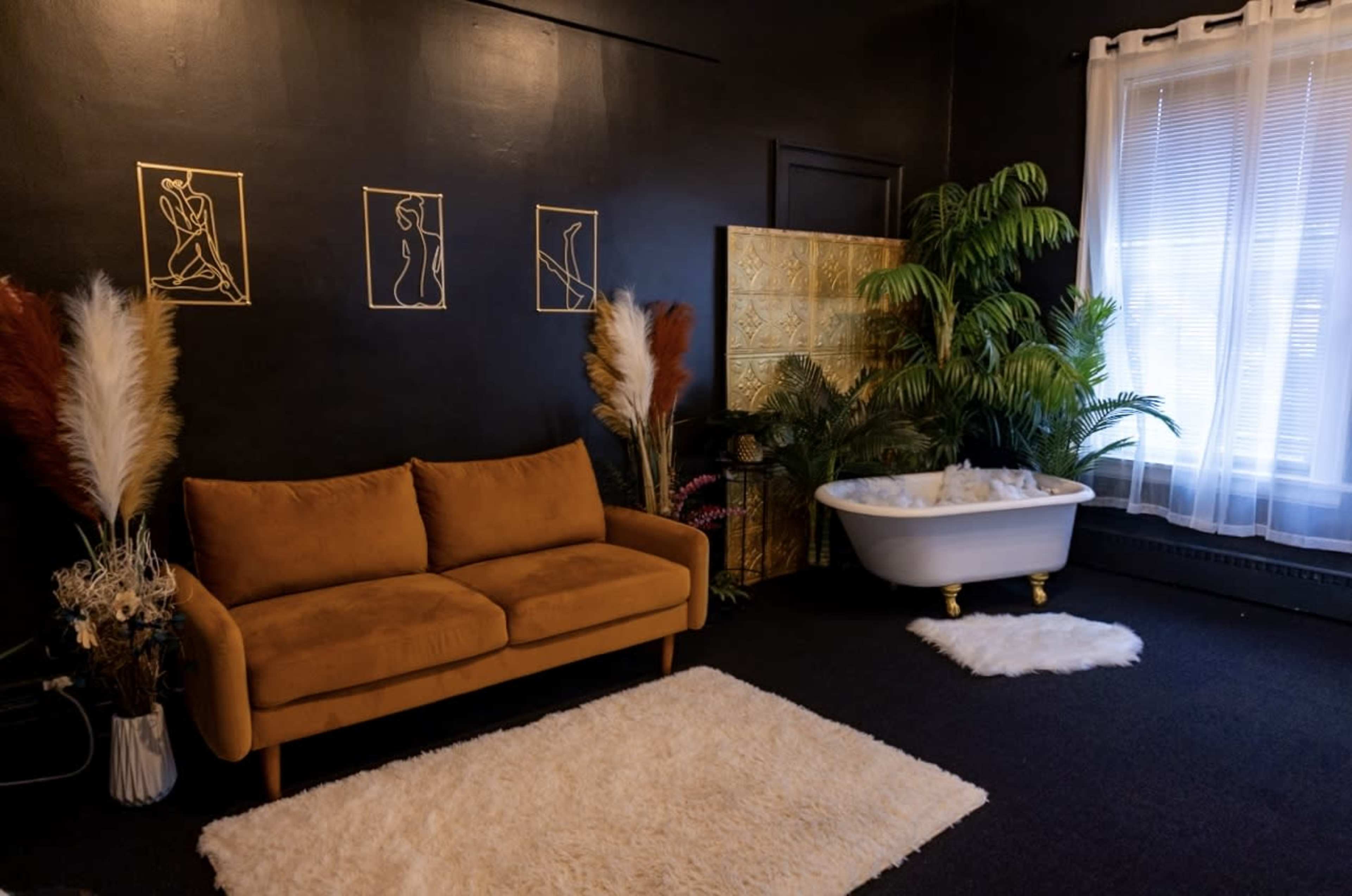 A dimly lit room with a brown couch, a white freestanding bathtub, decorative plants, and framed art on the walls.