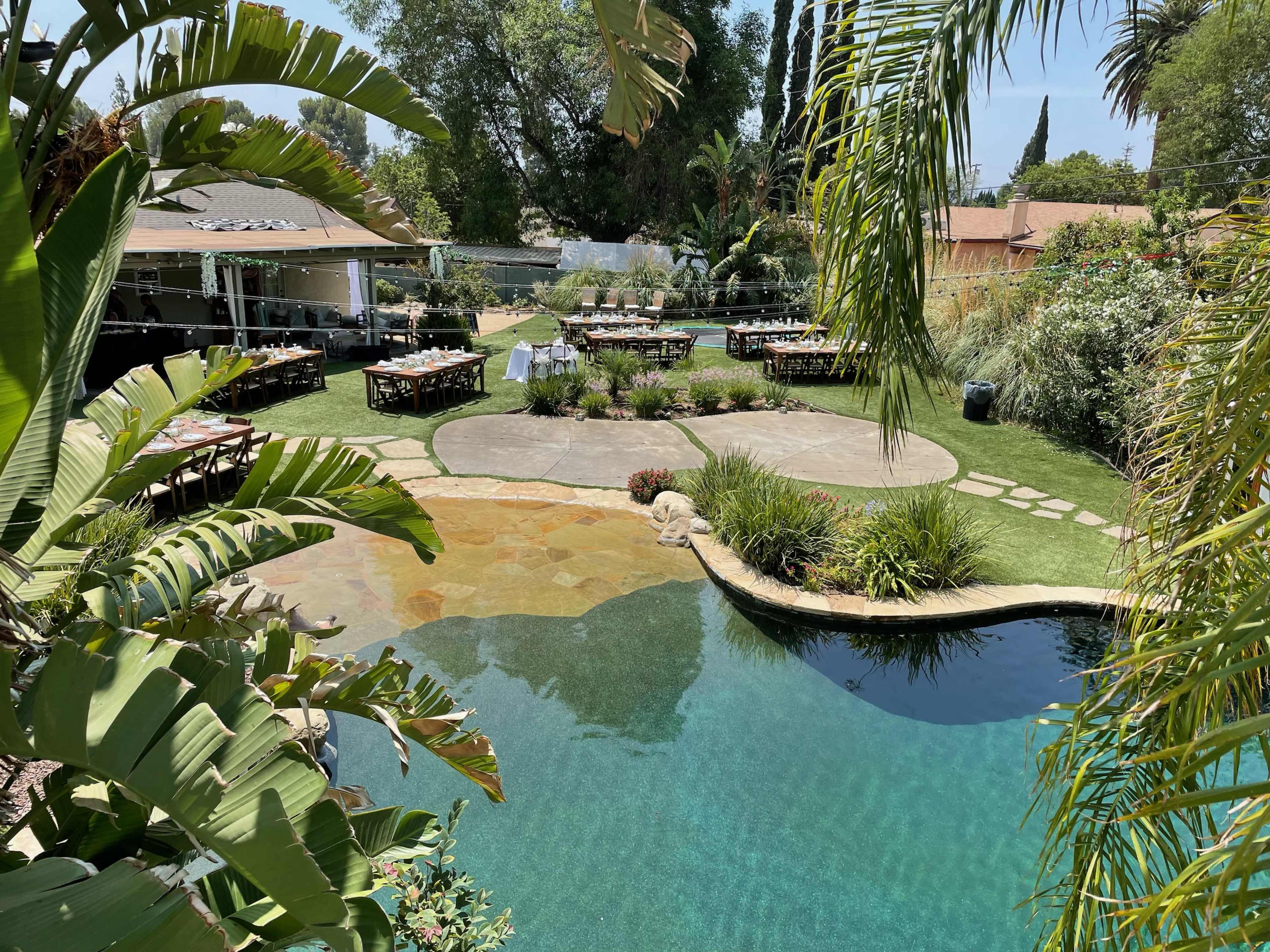 The image shows a landscaped backyard featuring a swimming pool, stone pathways, and multiple dining tables set up for an outdoor gathering.