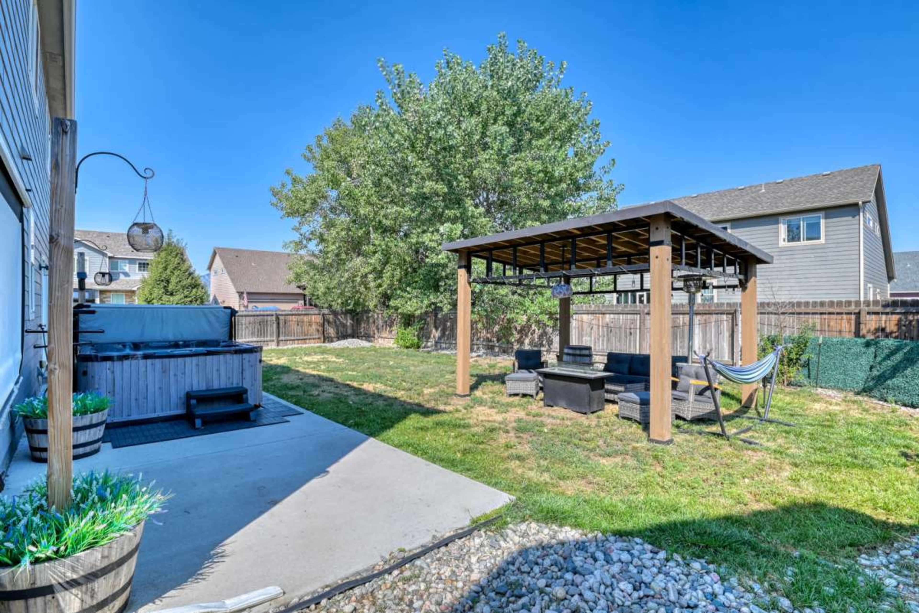 The image shows a backyard with a hot tub, a shaded seating area with a pergola, and a hammock, all surrounded by grass and a wooden fence.