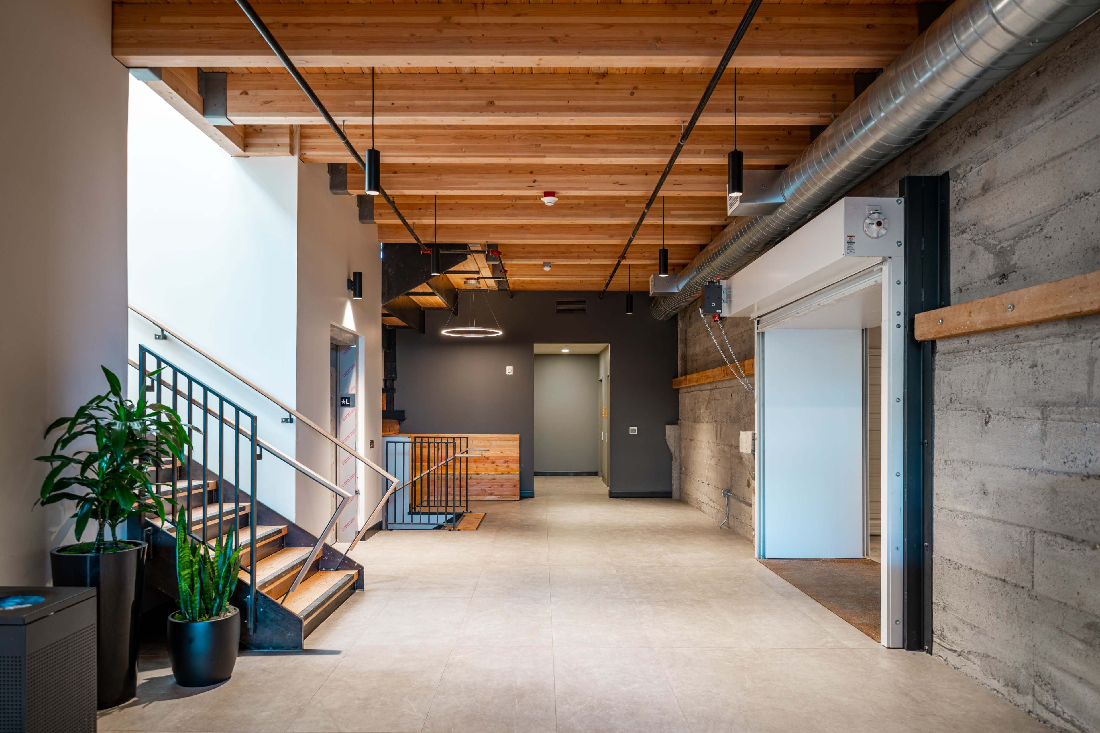 A modern, open lobby area featuring wooden beams, a staircase, and a potted plant against a backdrop of concrete walls.