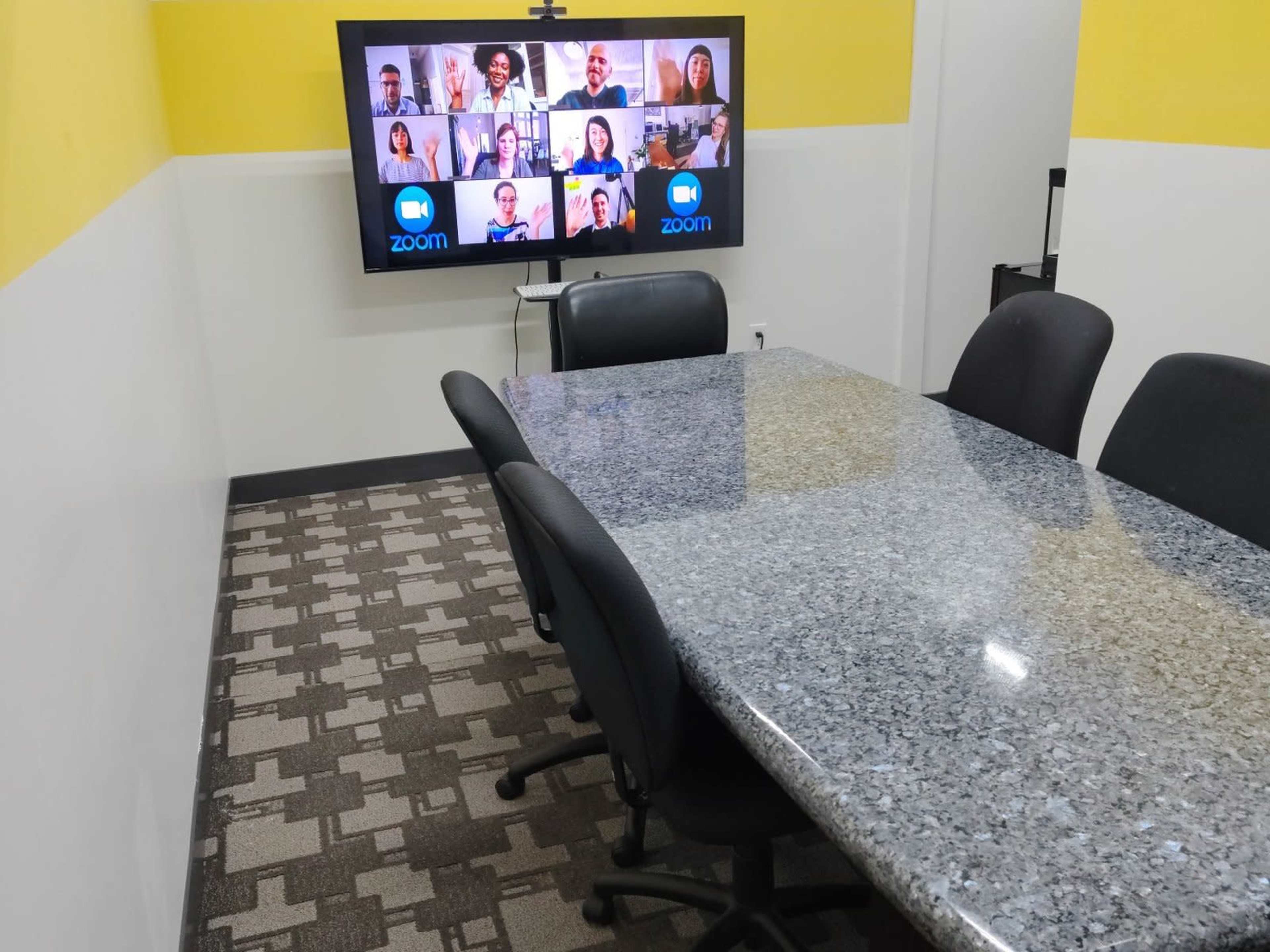 A conference room features a large table with black chairs and a wall-mounted screen displaying multiple participants in a video call.
