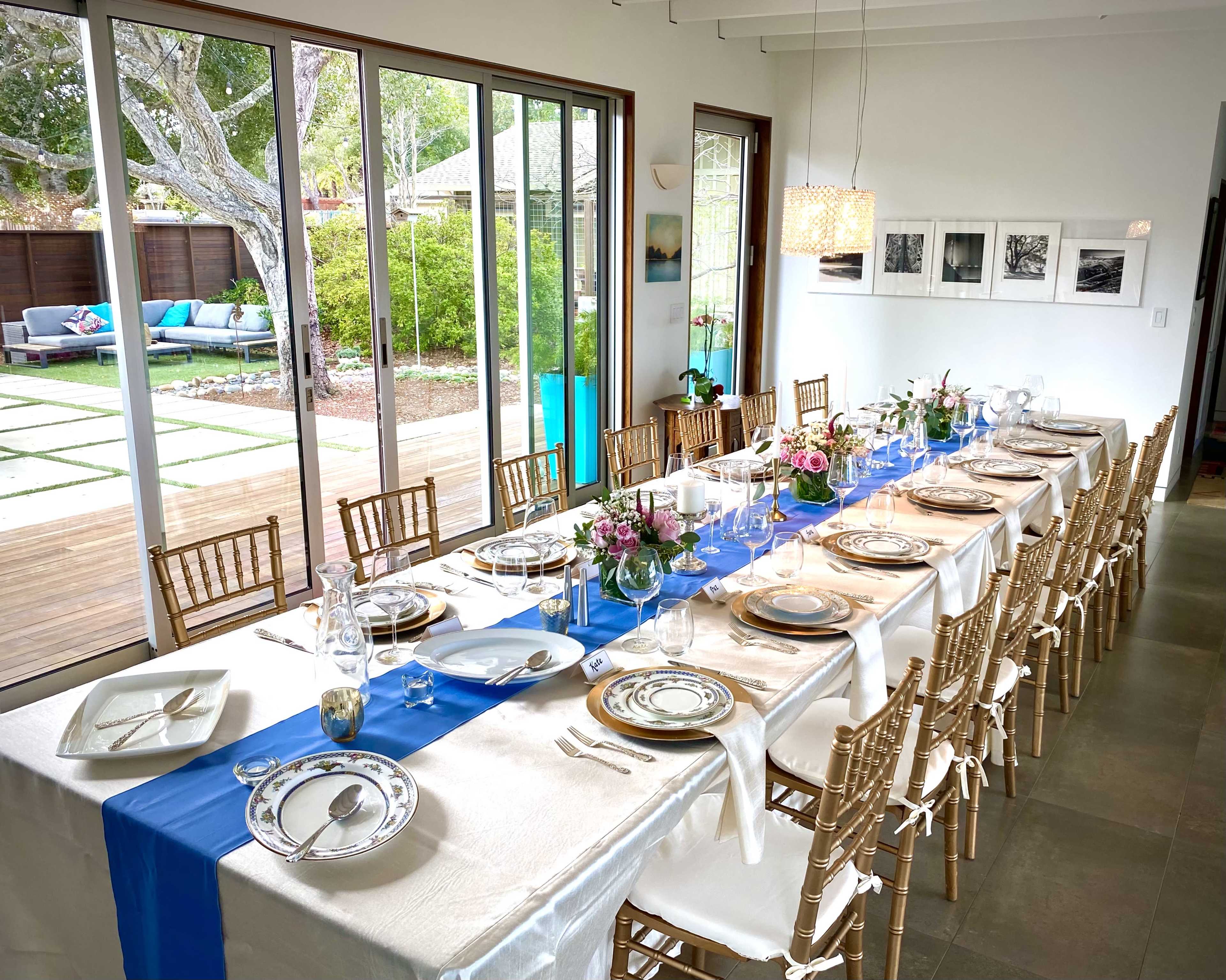 A long dining table set for a meal is arranged with elegant dishware and floral centerpieces, positioned in a bright room with large windows overlooking a garden.