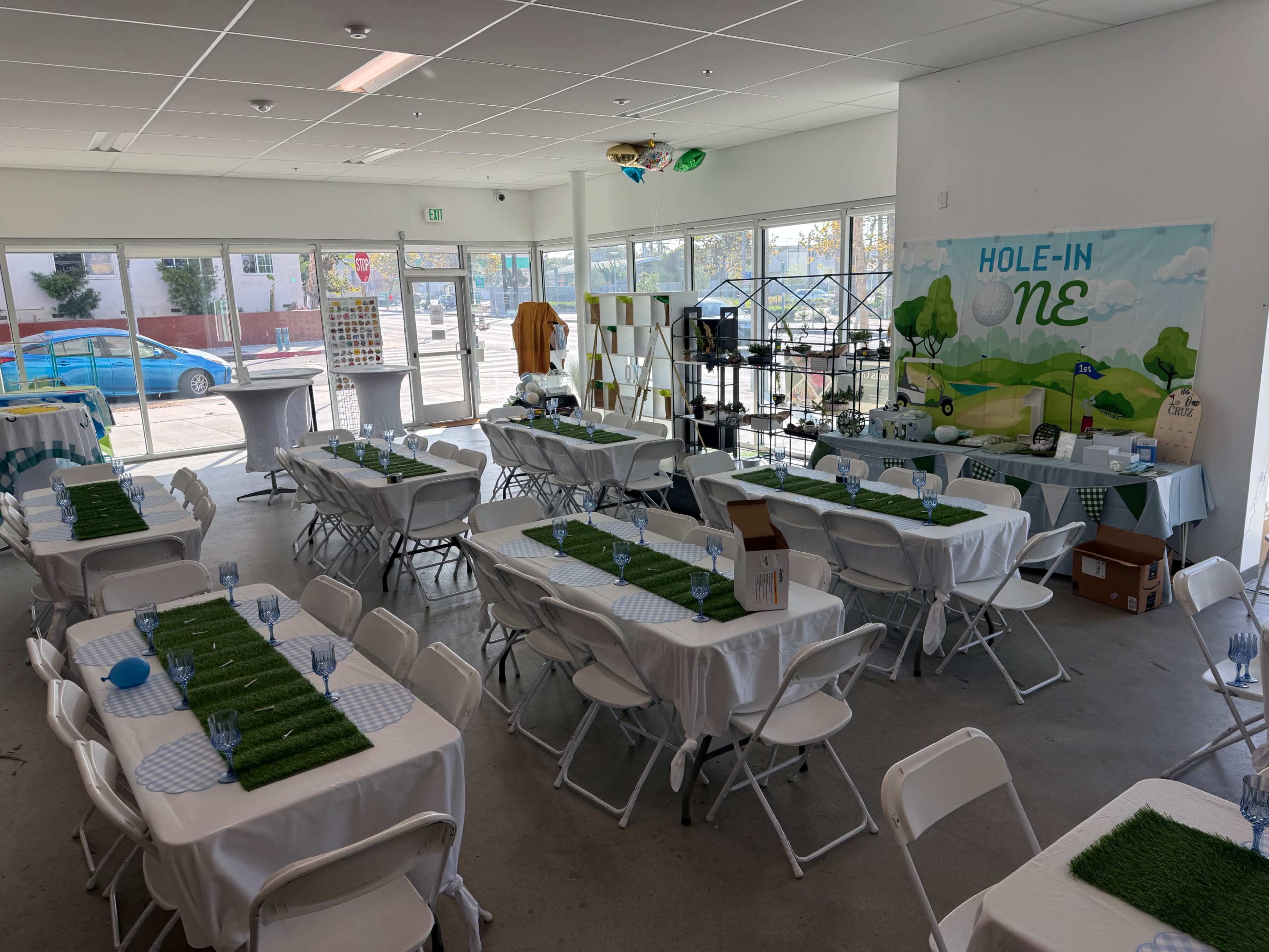 The image shows a spacious indoor event space set up with several tables covered in white tablecloths, each featuring green grass-like decorations, and a large mural depicting a landscape in the background.