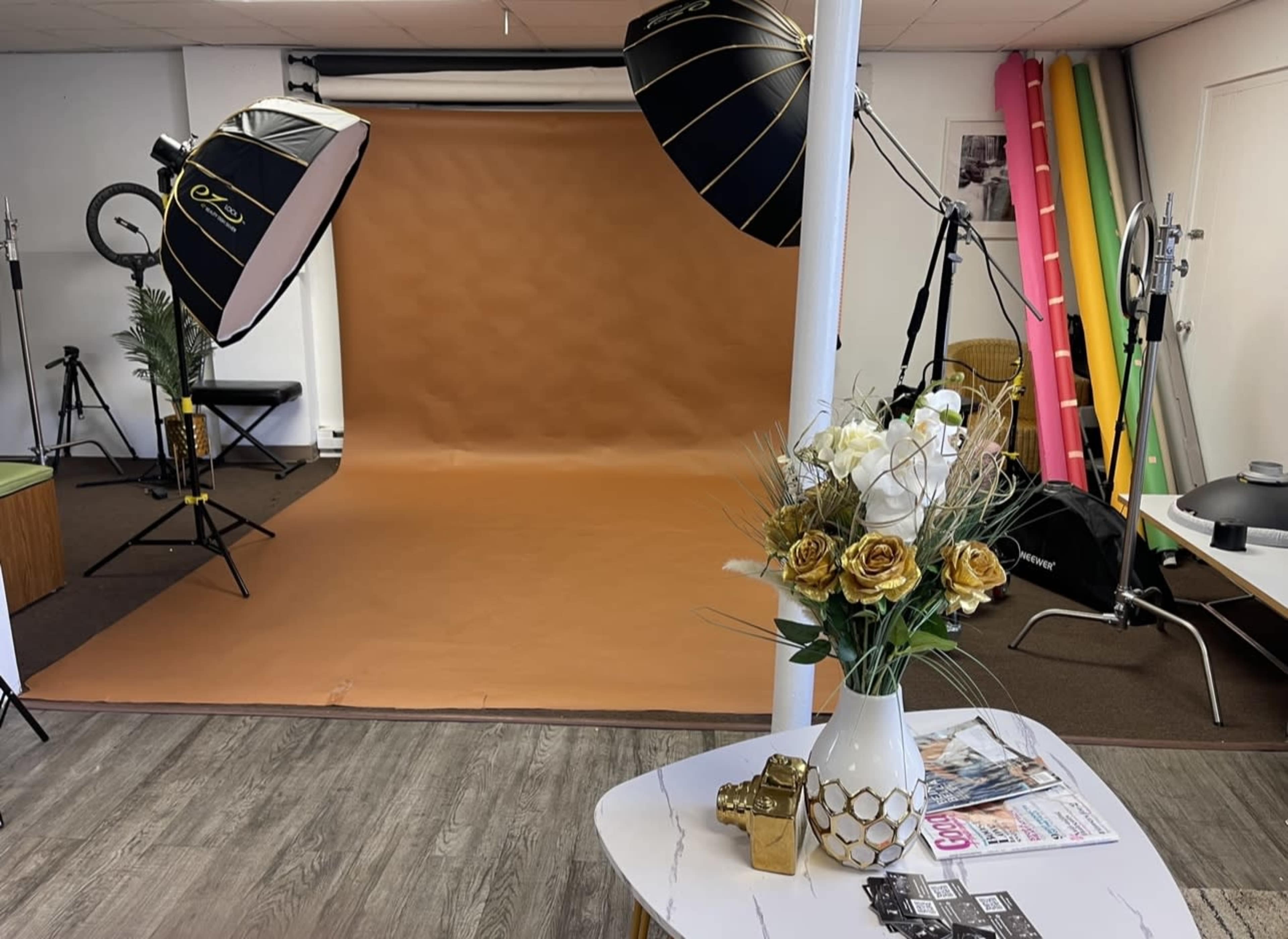 A photography studio featuring a brown backdrop, two large softbox lights, a decorative flower arrangement on a table, and various photography equipment in the background.