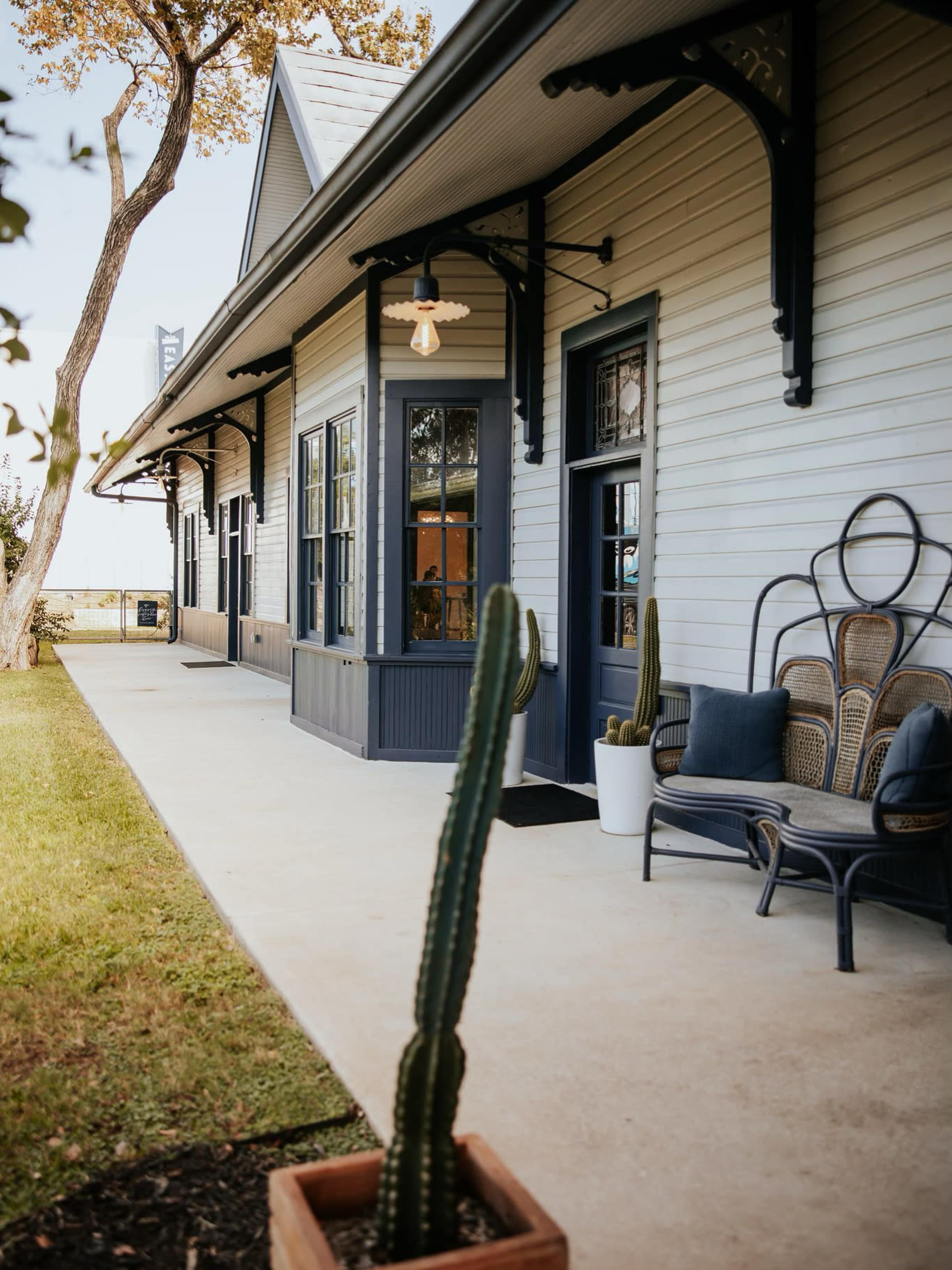 The image shows a light-colored house with a covered porch, featuring a seating area and potted cacti along the walkway.
