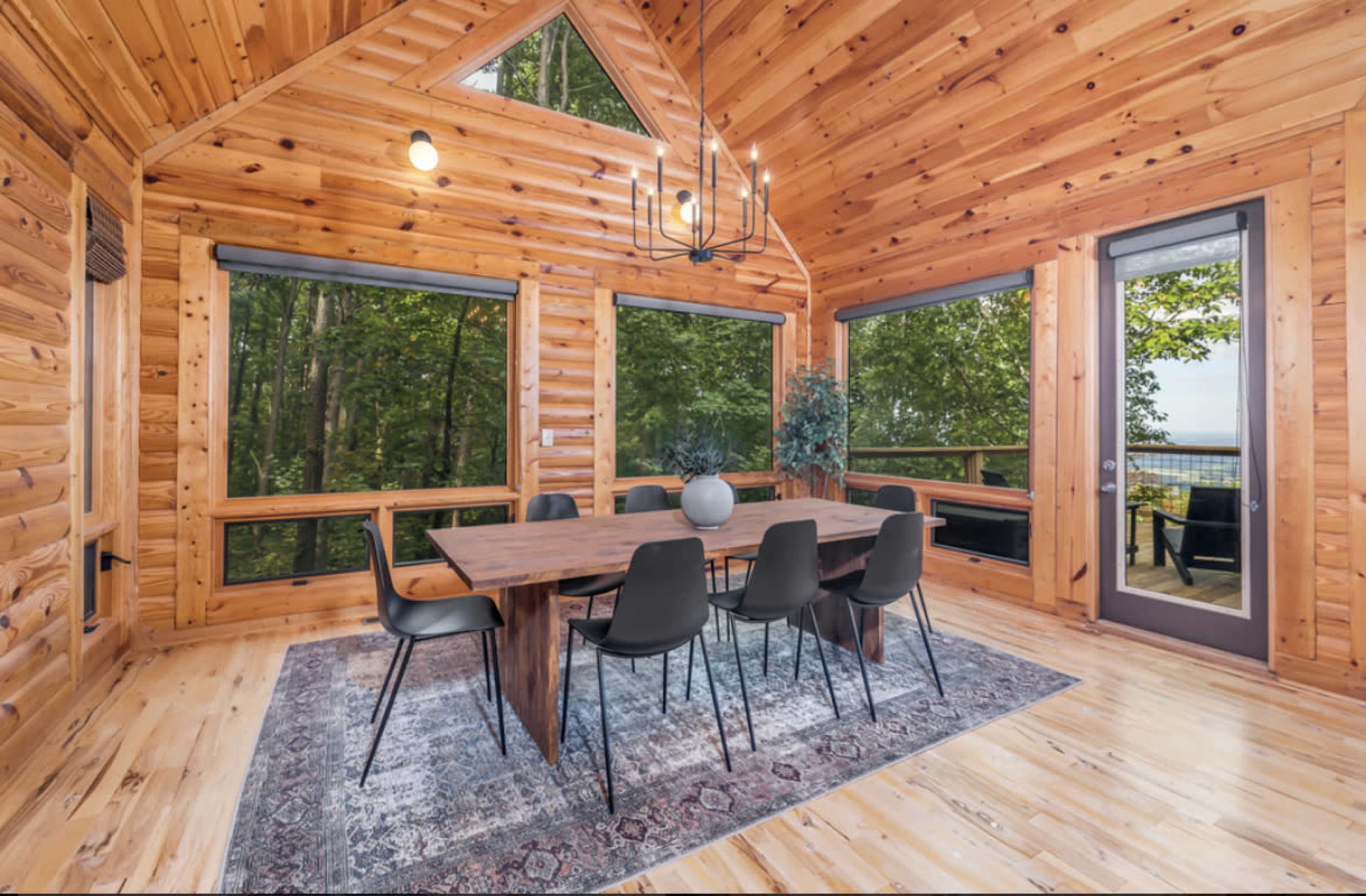 A wooden dining area features a large table surrounded by six chairs, with large windows overlooking a forested outdoor space.