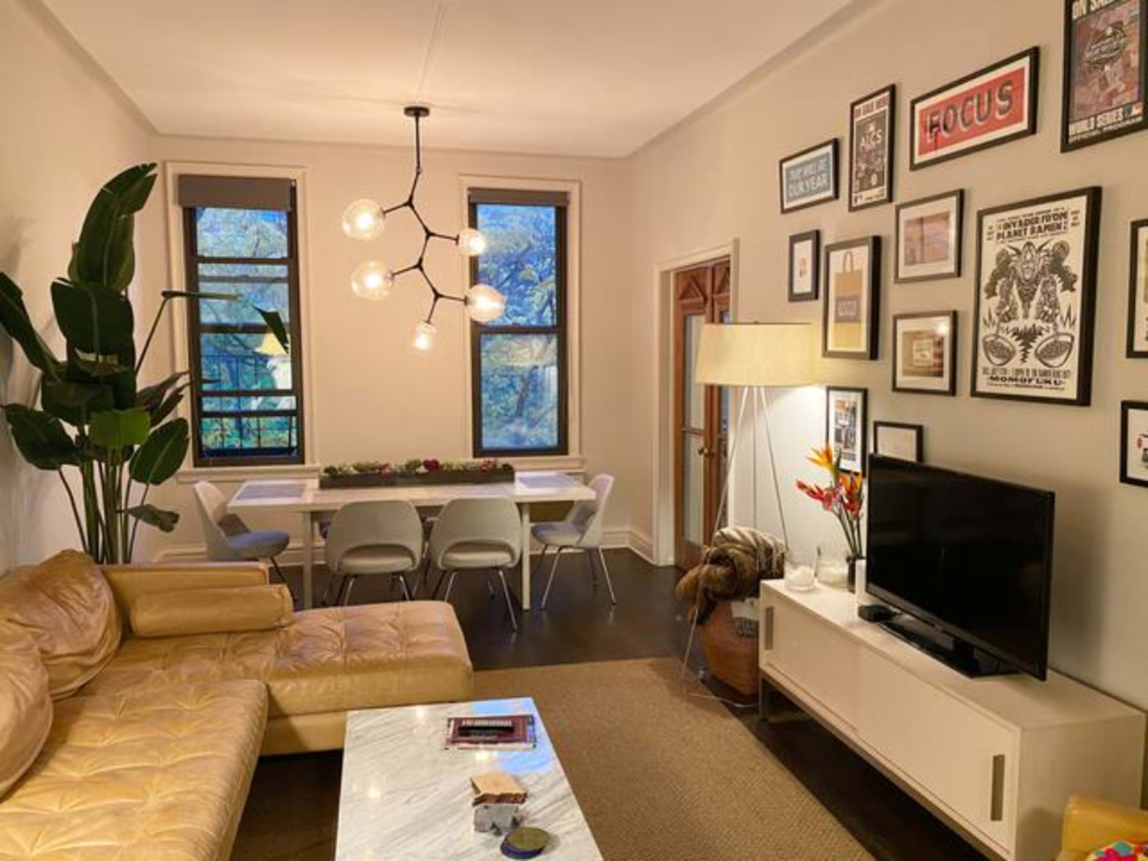 A living room with a tan sectional sofa, a white coffee table, a dining area with modern chairs, and various framed artworks on the walls.