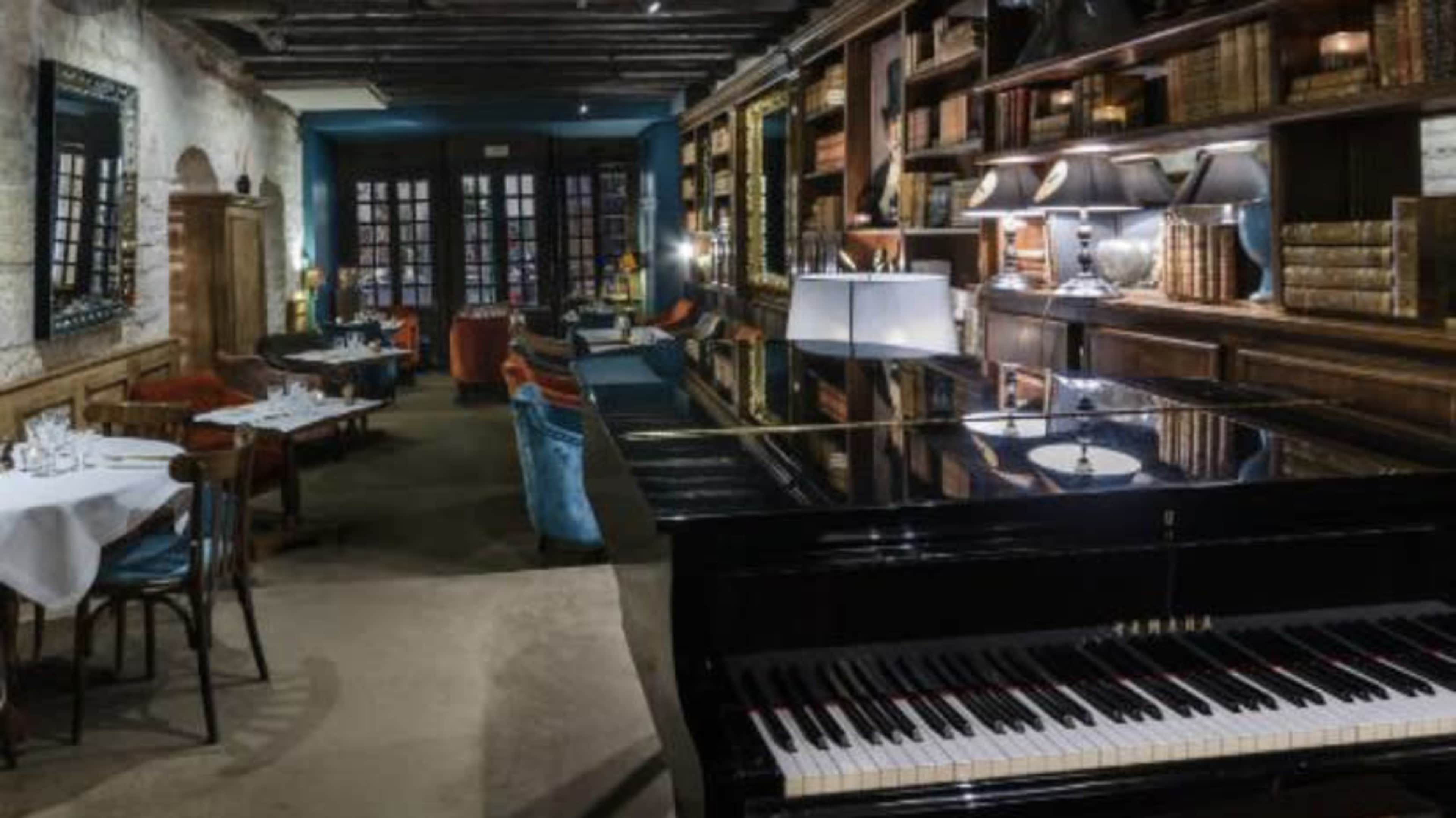 An elegant dining area with wooden shelves filled with books, a grand piano in the foreground, and tables set for dining in a softly lit atmosphere.