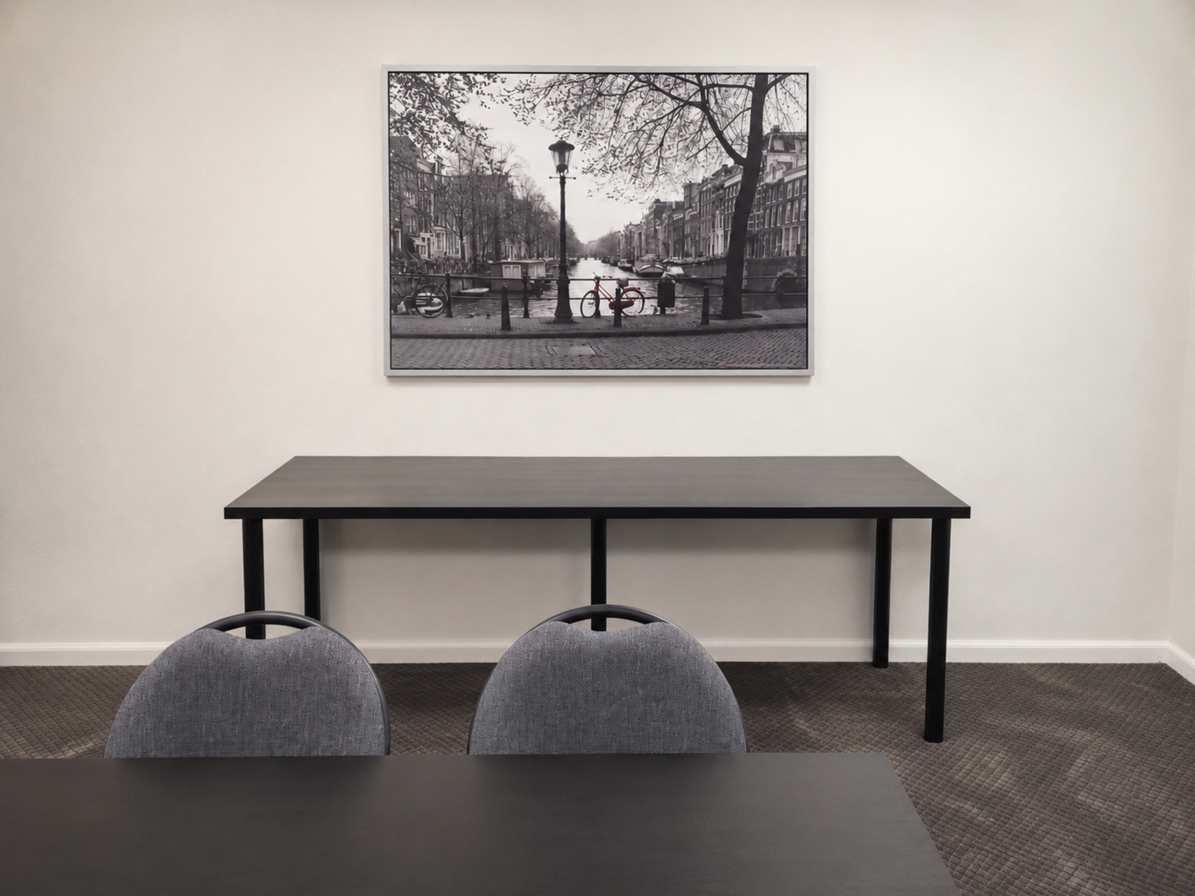 A simple meeting room features a long black table with two gray chairs and a black-and-white photograph of a canal scene on the wall.
