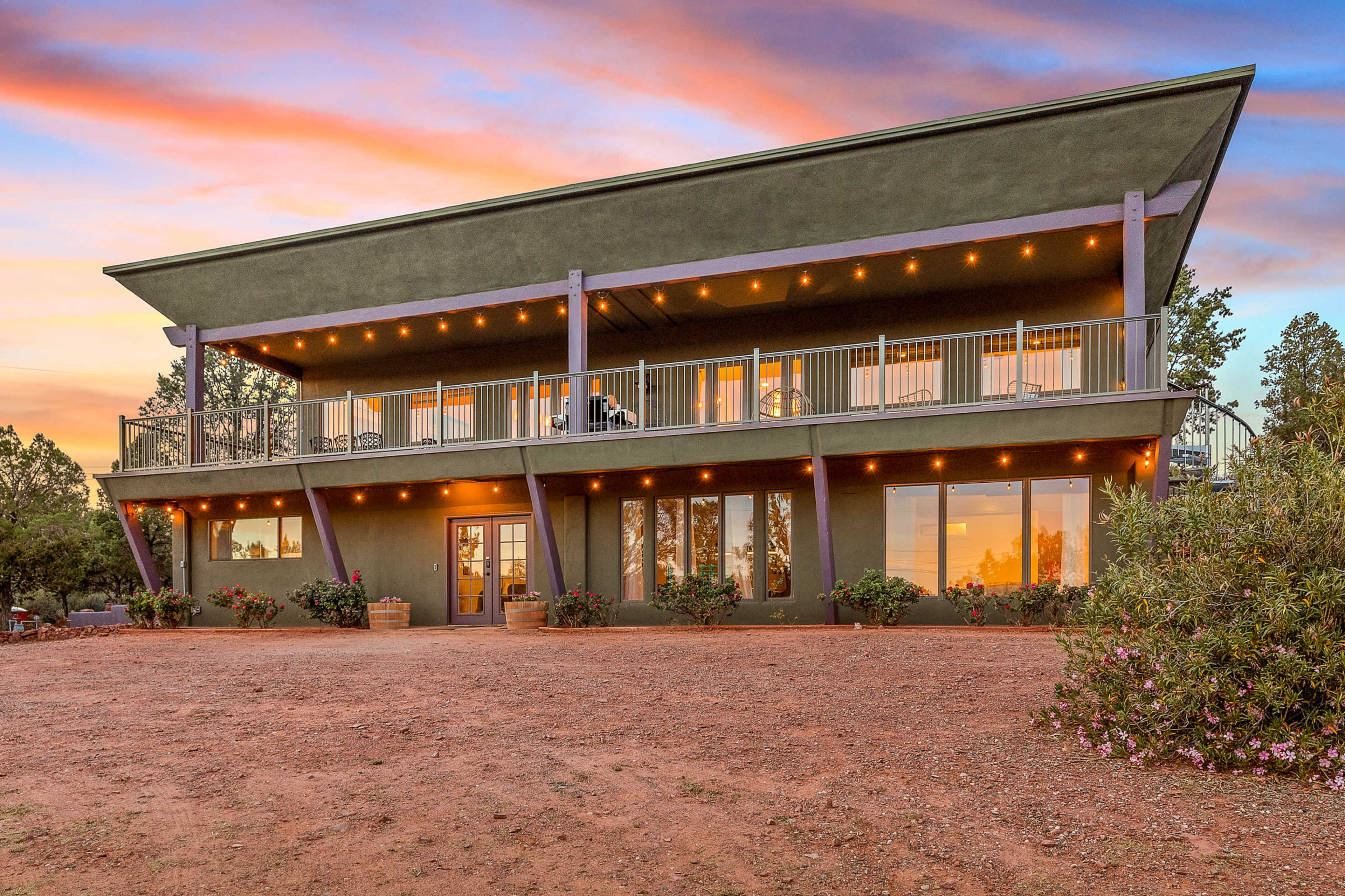A modern two-story green house with a balcony, surrounded by gravel and landscaped plants, against a colorful sunset sky.
