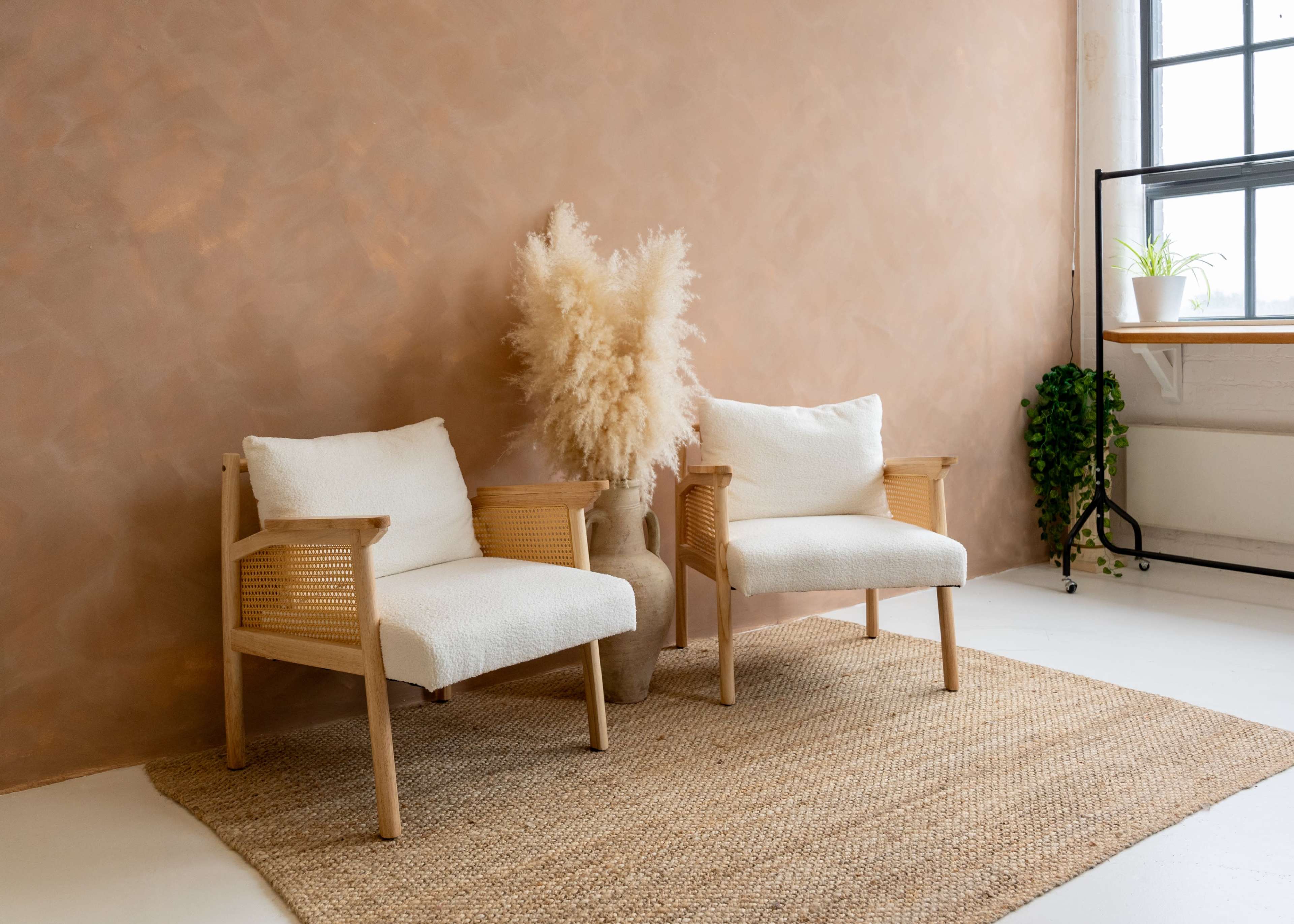 The image features two white upholstered chairs with wooden frames positioned on a woven rug in front of a textured brown wall, alongside a large vase filled with pampas grass.