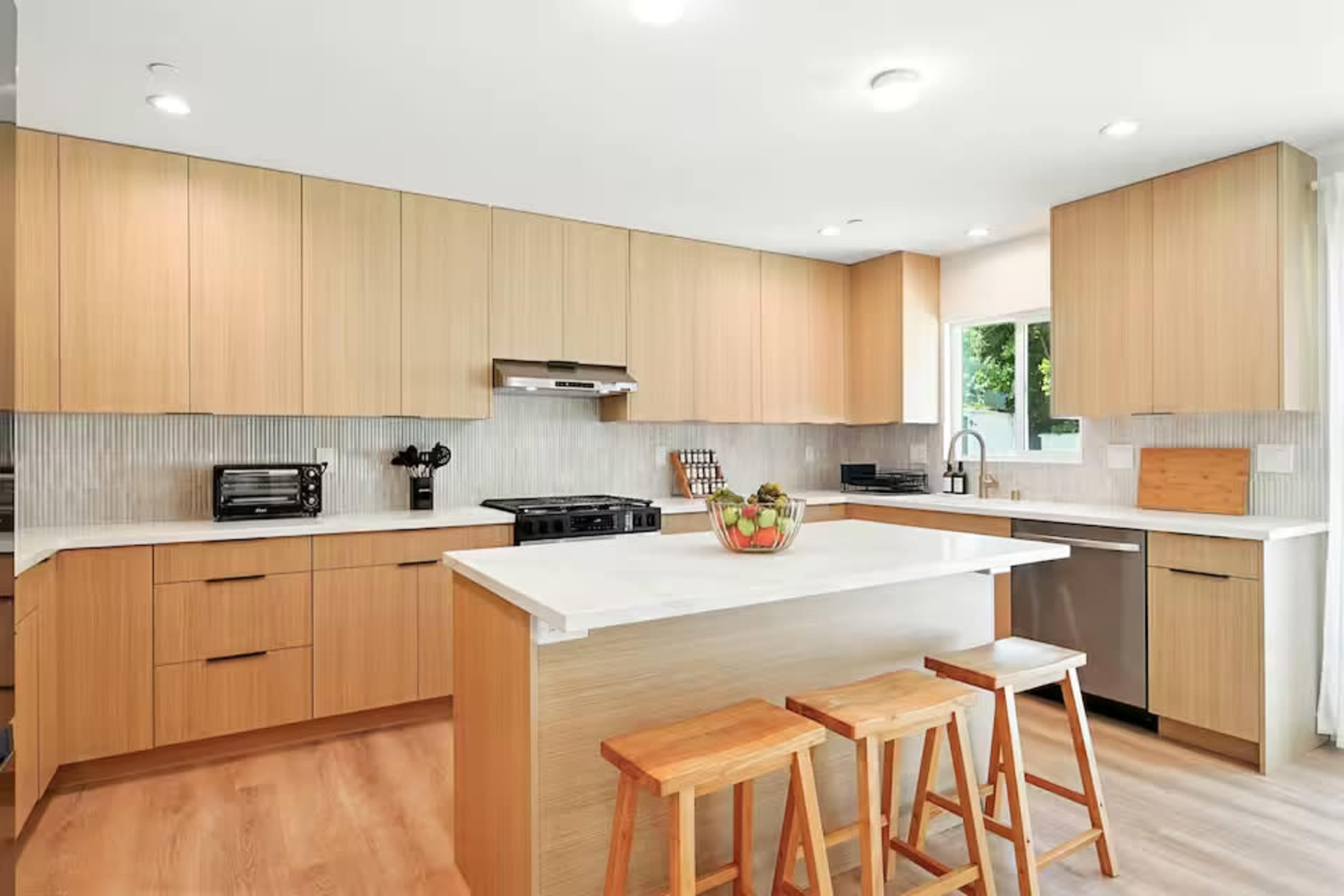 The image shows a modern kitchen with wooden cabinets, a central island with three stools, and stainless steel appliances.