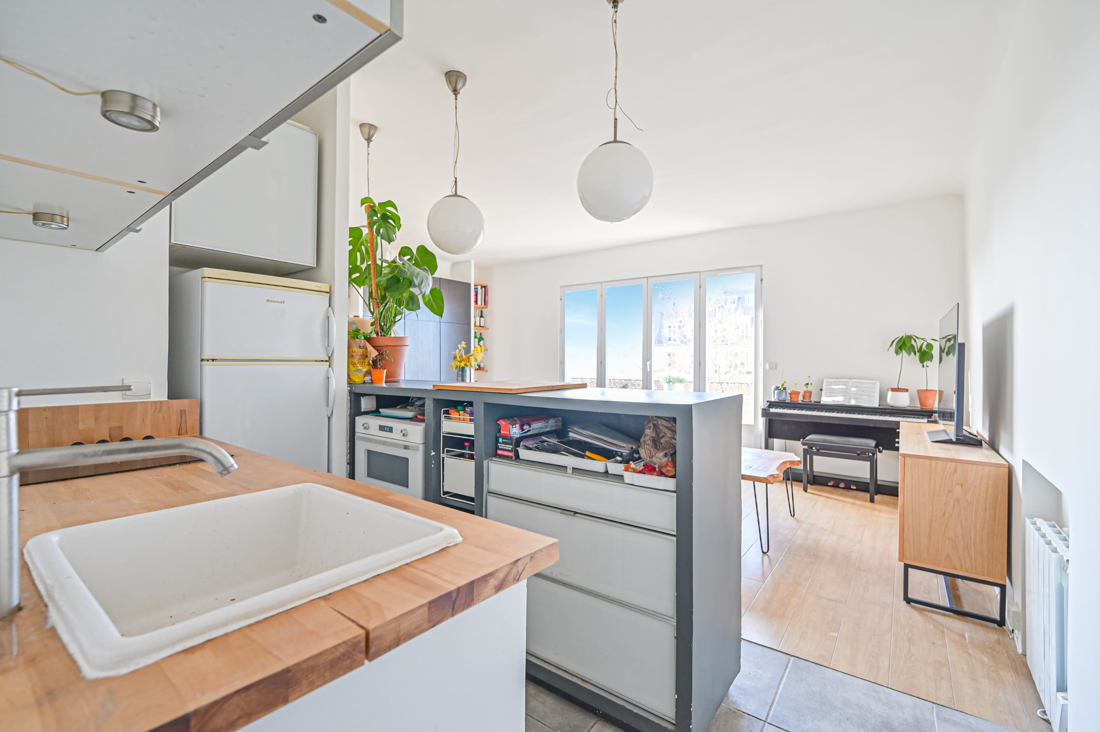 The image shows a modern kitchen with an island and a clear view into a bright living area that features a piano and a desk.