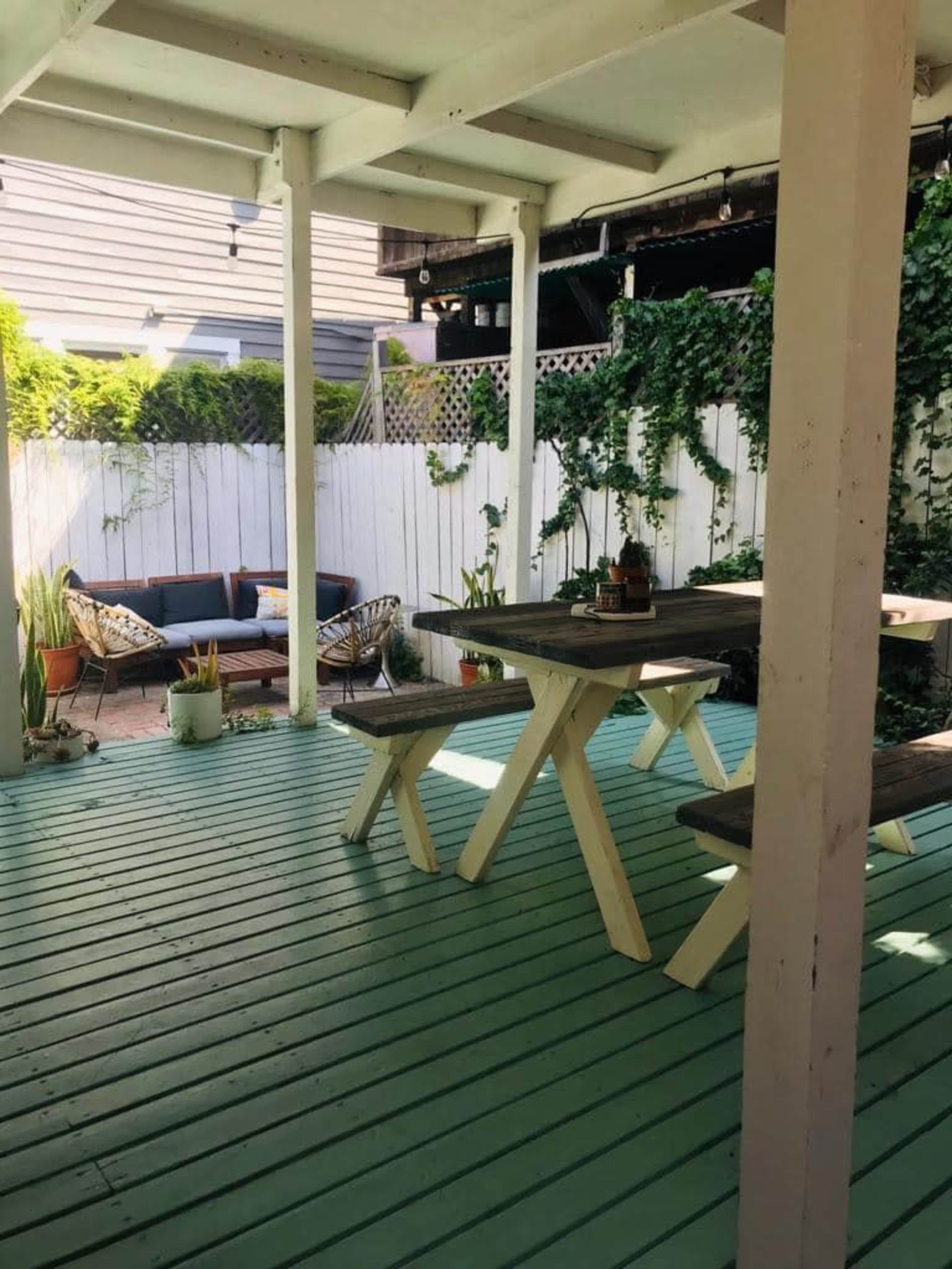 A covered porch with a green wooden floor, a dining table, and a seating area surrounded by plants and a white fence.