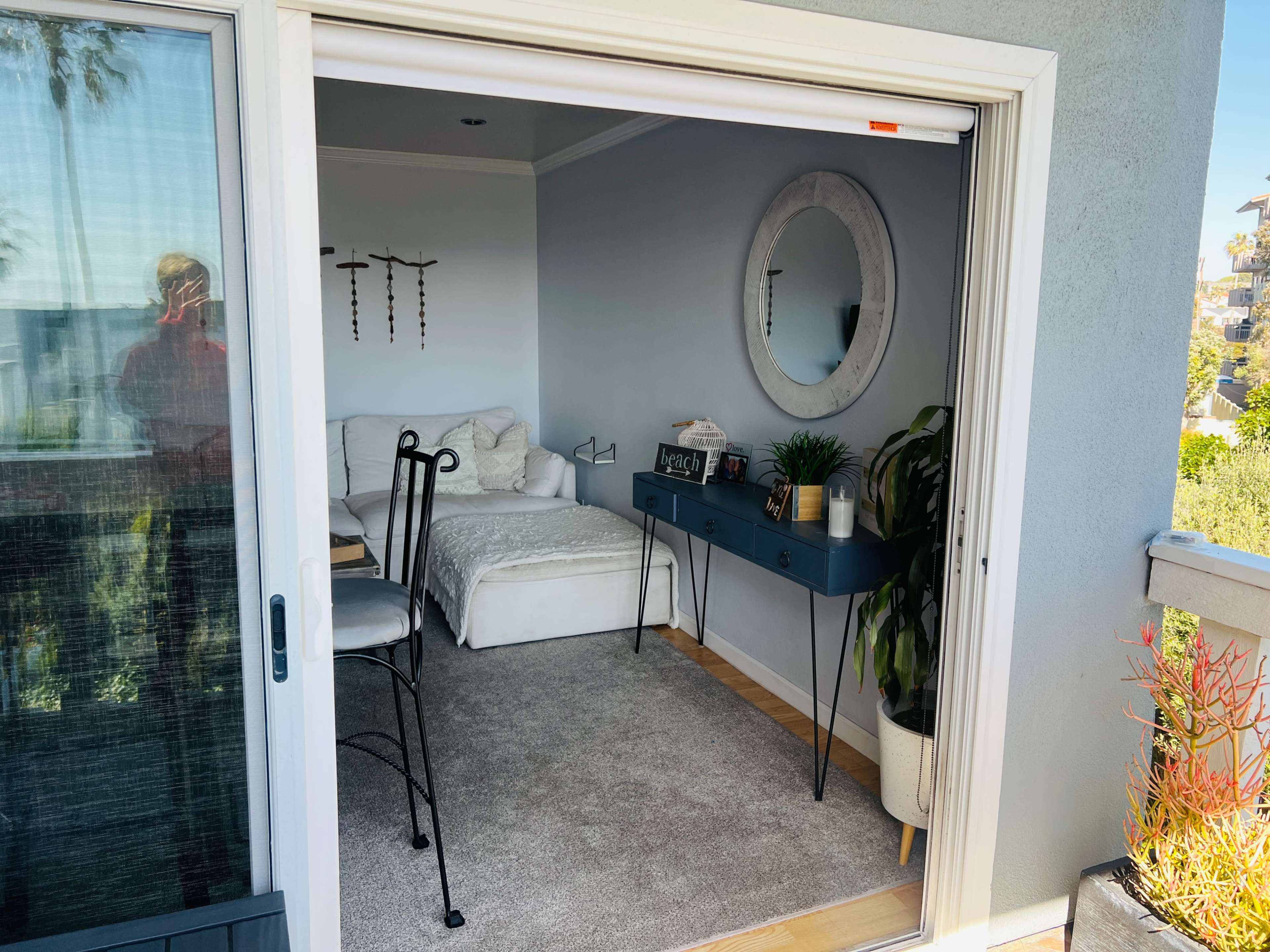 A small, minimalist room with a bed, a desk, and a large round mirror, seen through an open sliding glass door.