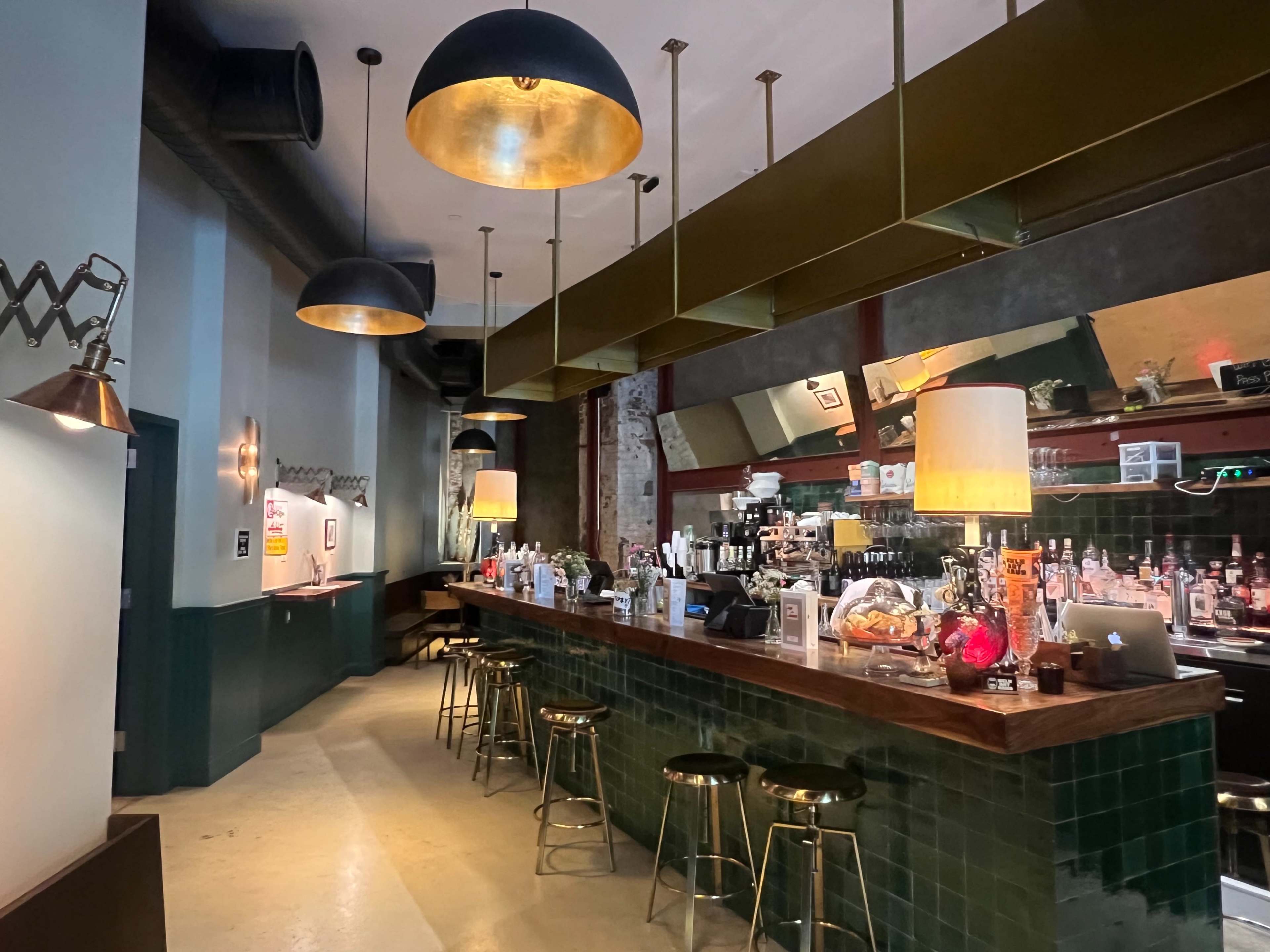 The image shows a bar area with a long wooden counter, shelves displaying various bottles, and overhead pendant lights casting ambient illumination.
