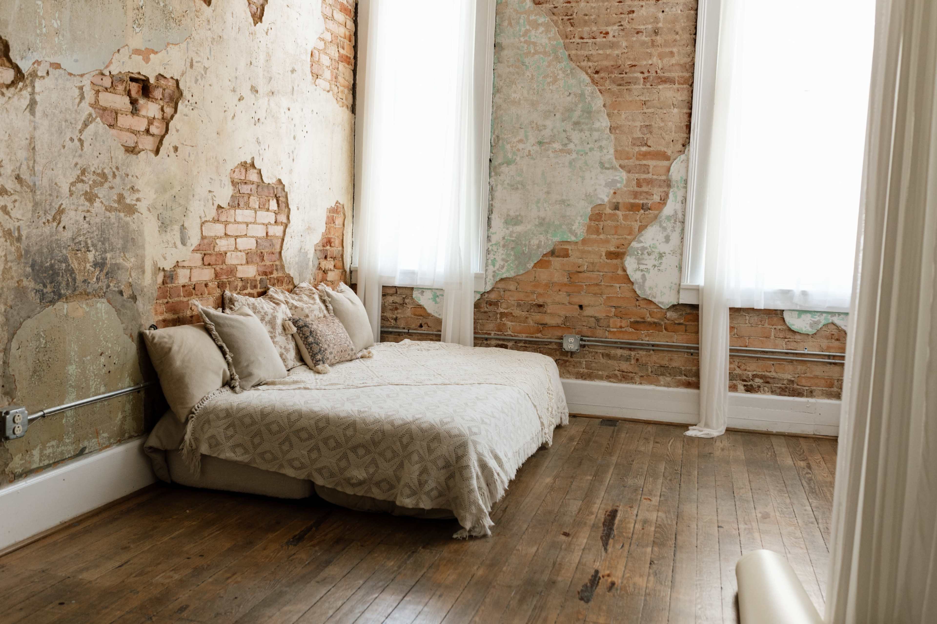 The image shows a simple bedroom featuring a low, unmade bed with a light-colored quilt against a wall with exposed brick and peeling paint.