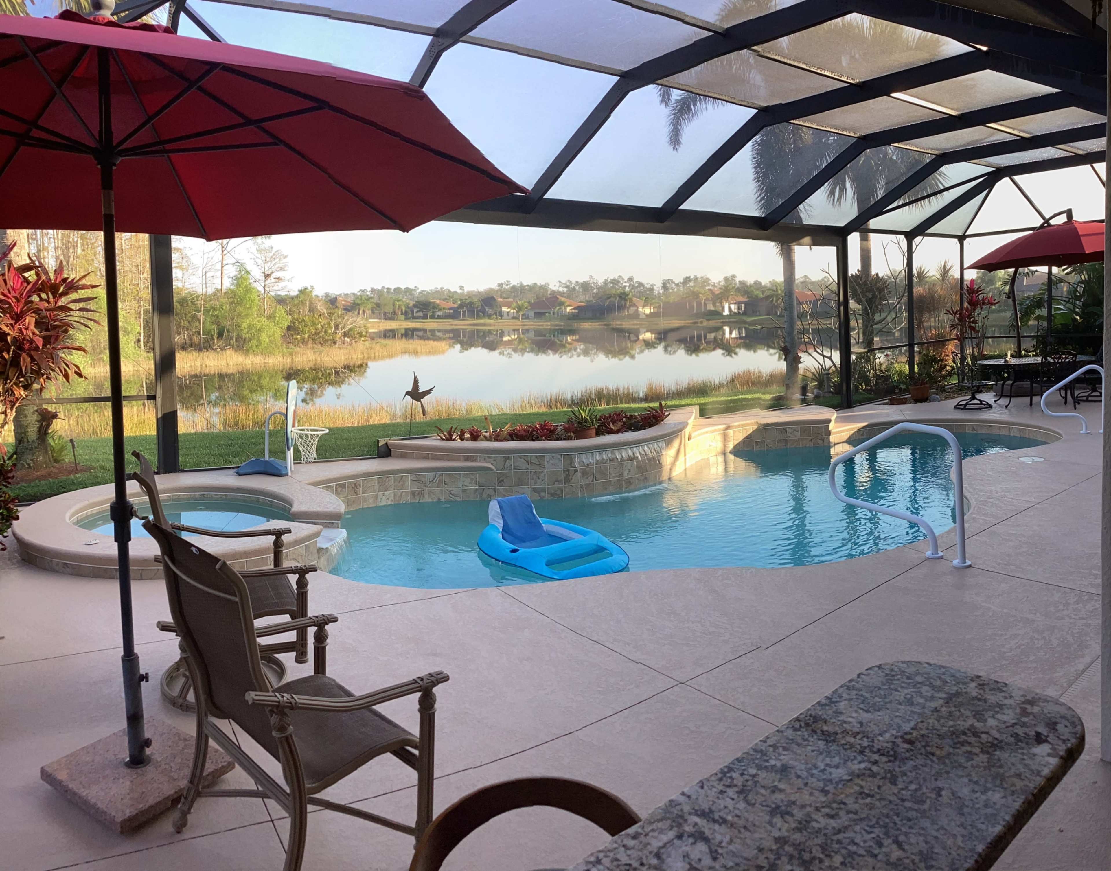 A pool area enclosed by a screen, featuring a hot tub, a blue float, and shaded seating, overlooking a calm body of water surrounded by greenery.
