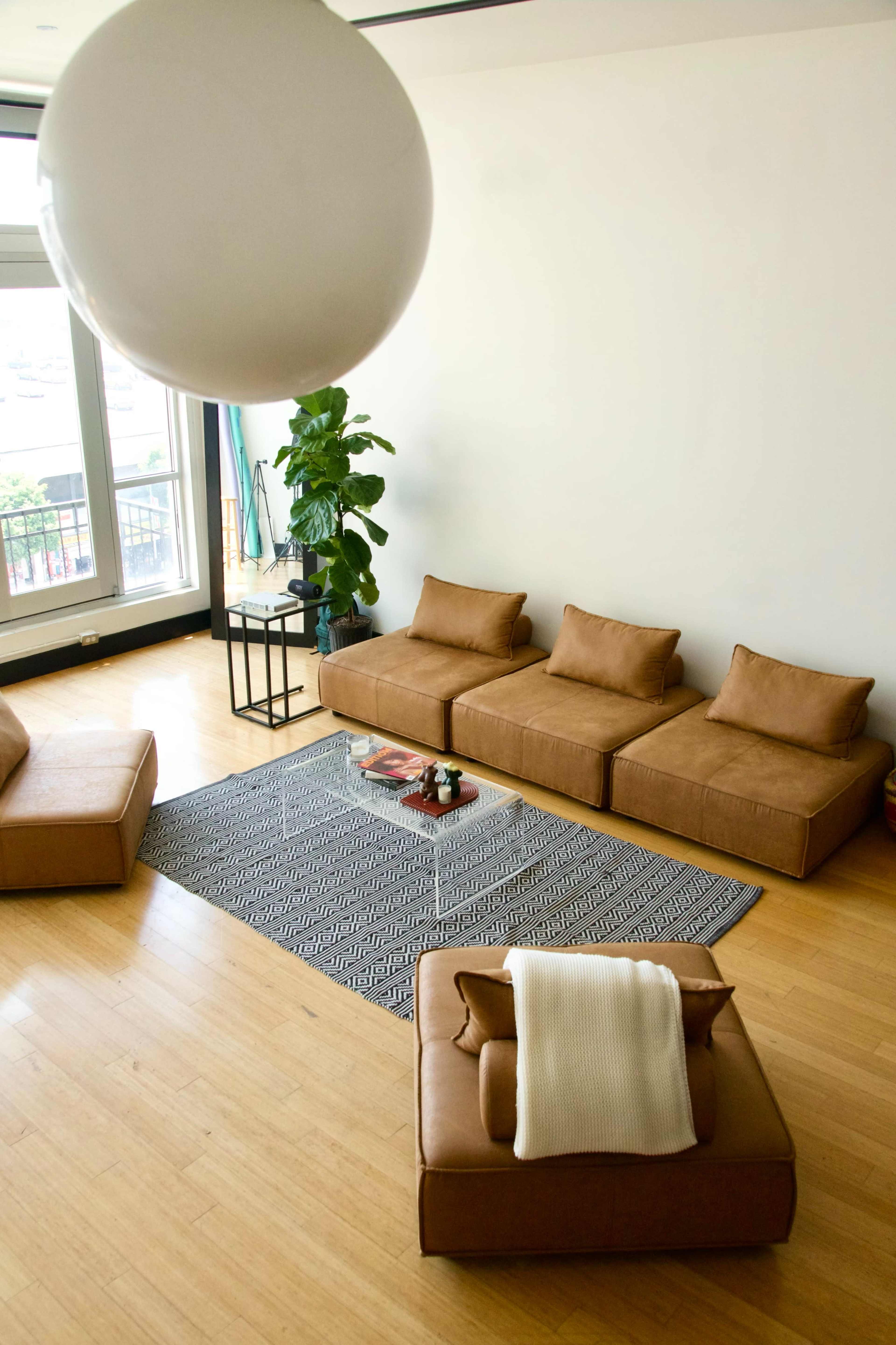 A modern living room with a geometric patterned rug, brown seating arrangements, a round pendant light, and a potted plant.