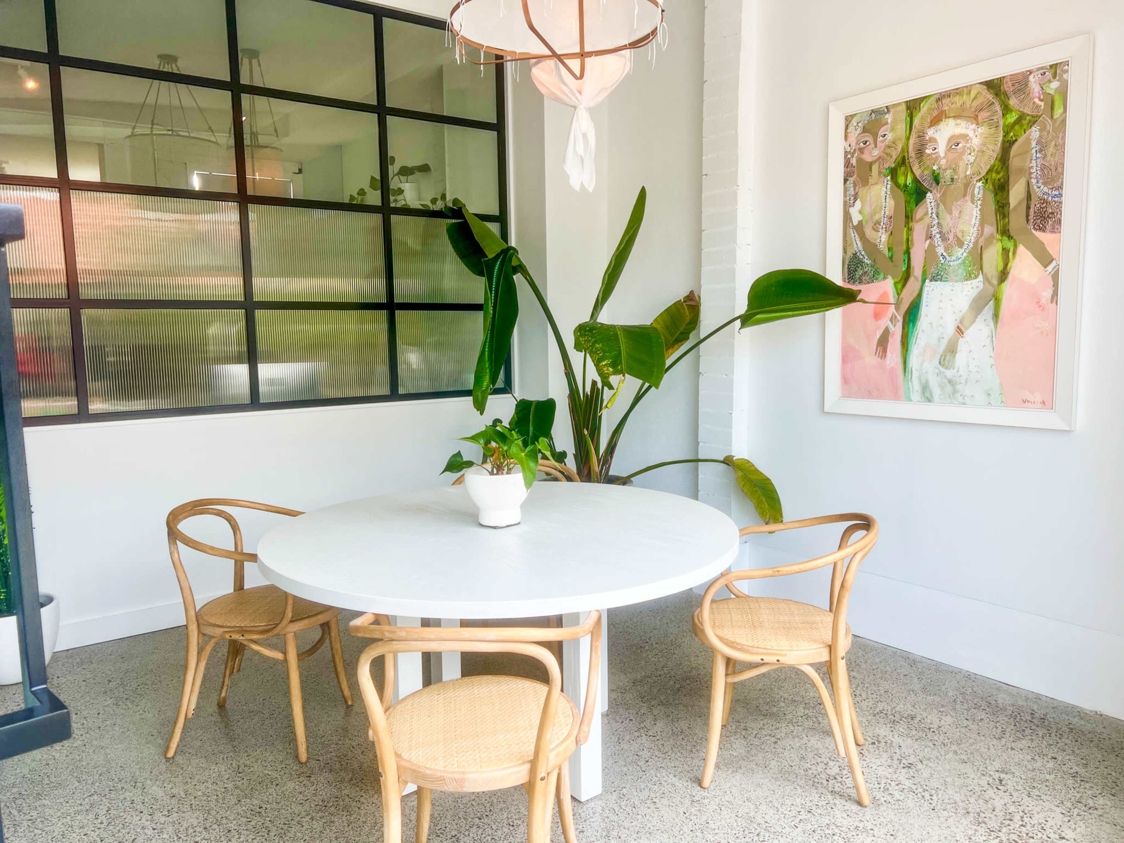 A round white table with four woven chairs is positioned in a bright room featuring a large window and a potted plant.