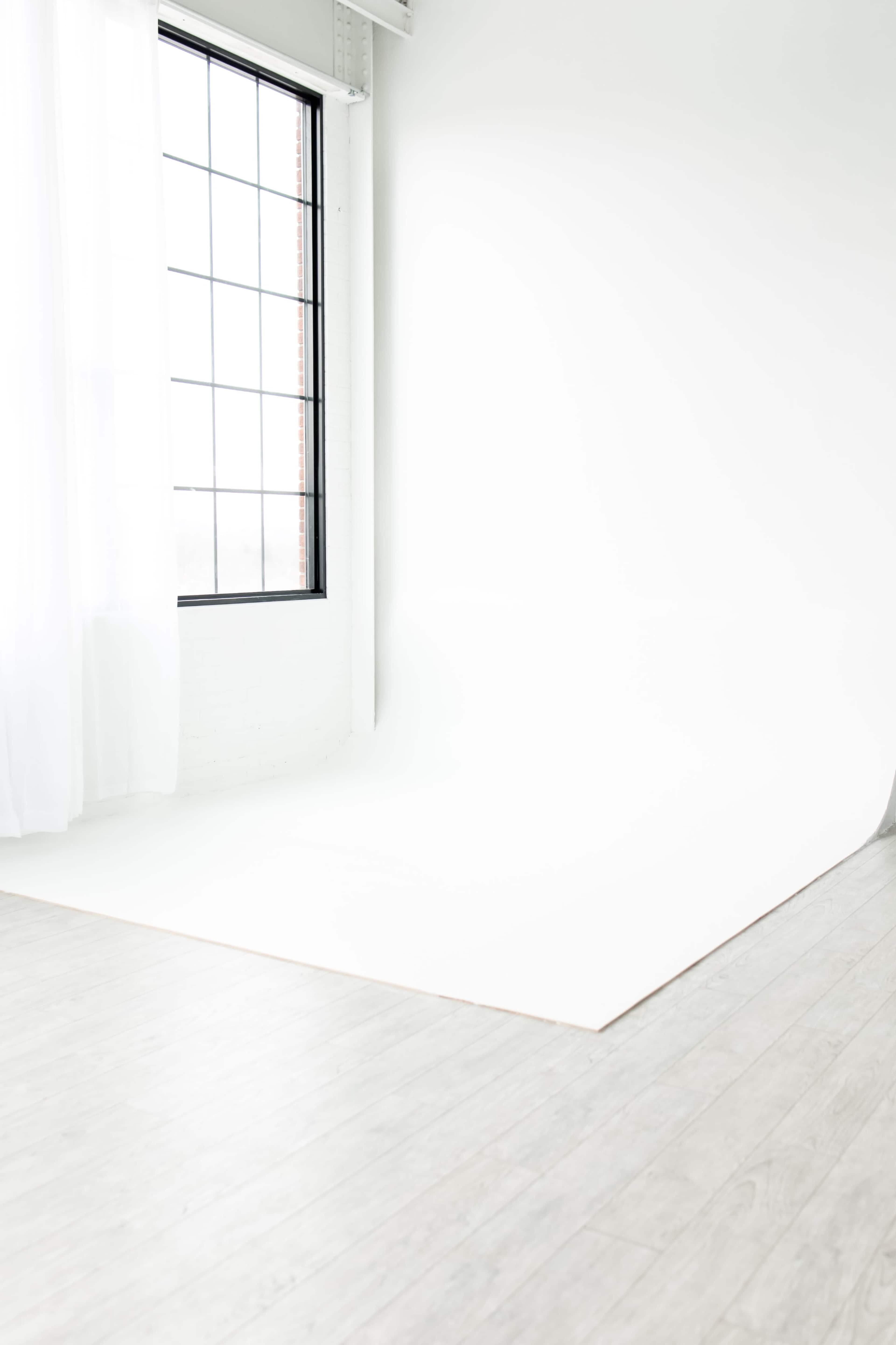 The image shows a bright, minimalist interior space with a large window and a white backdrop on the floor.