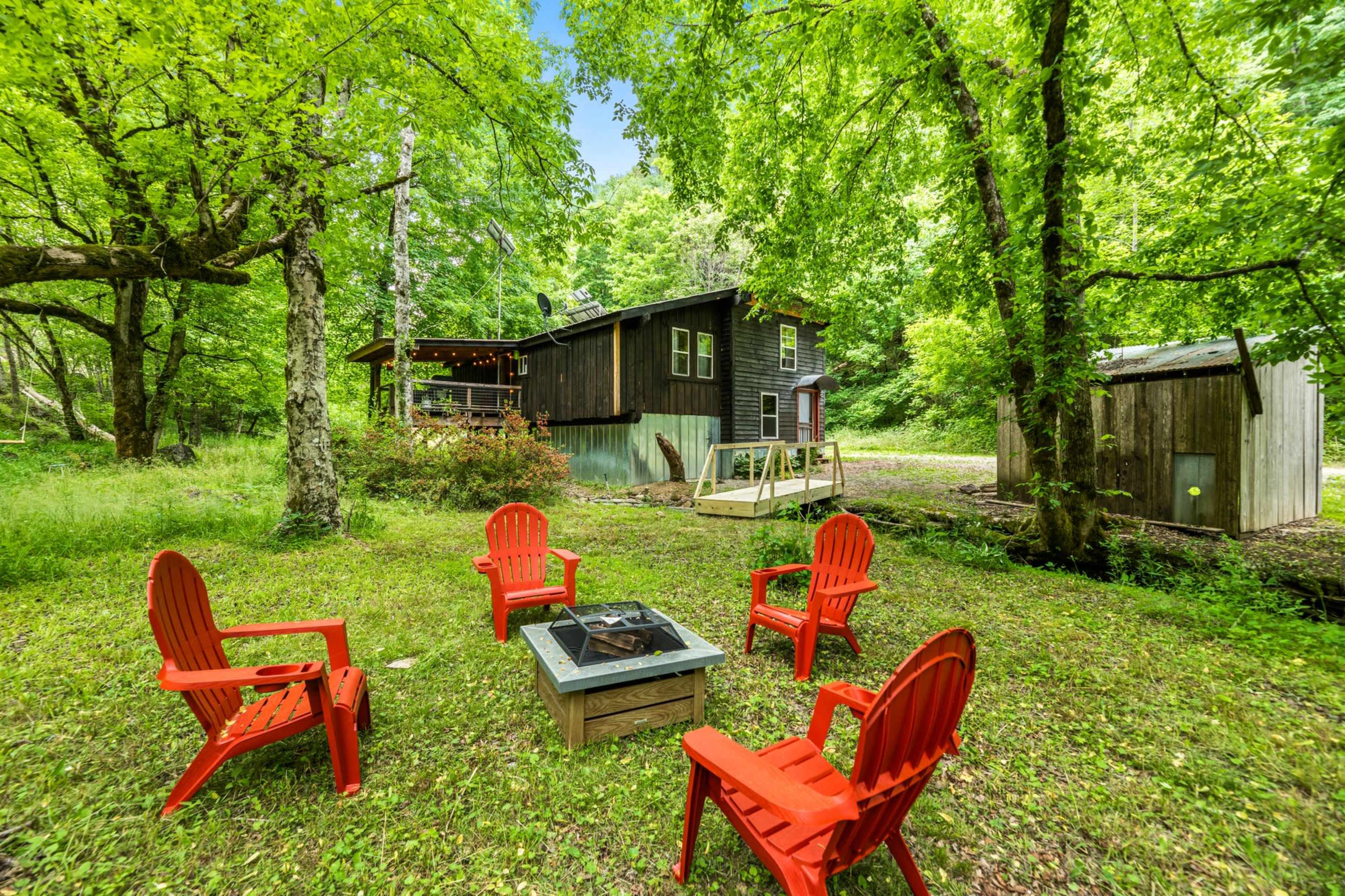 A group of red chairs surrounds a fire pit in a grassy area near a two-story cabin, surrounded by trees and greenery.