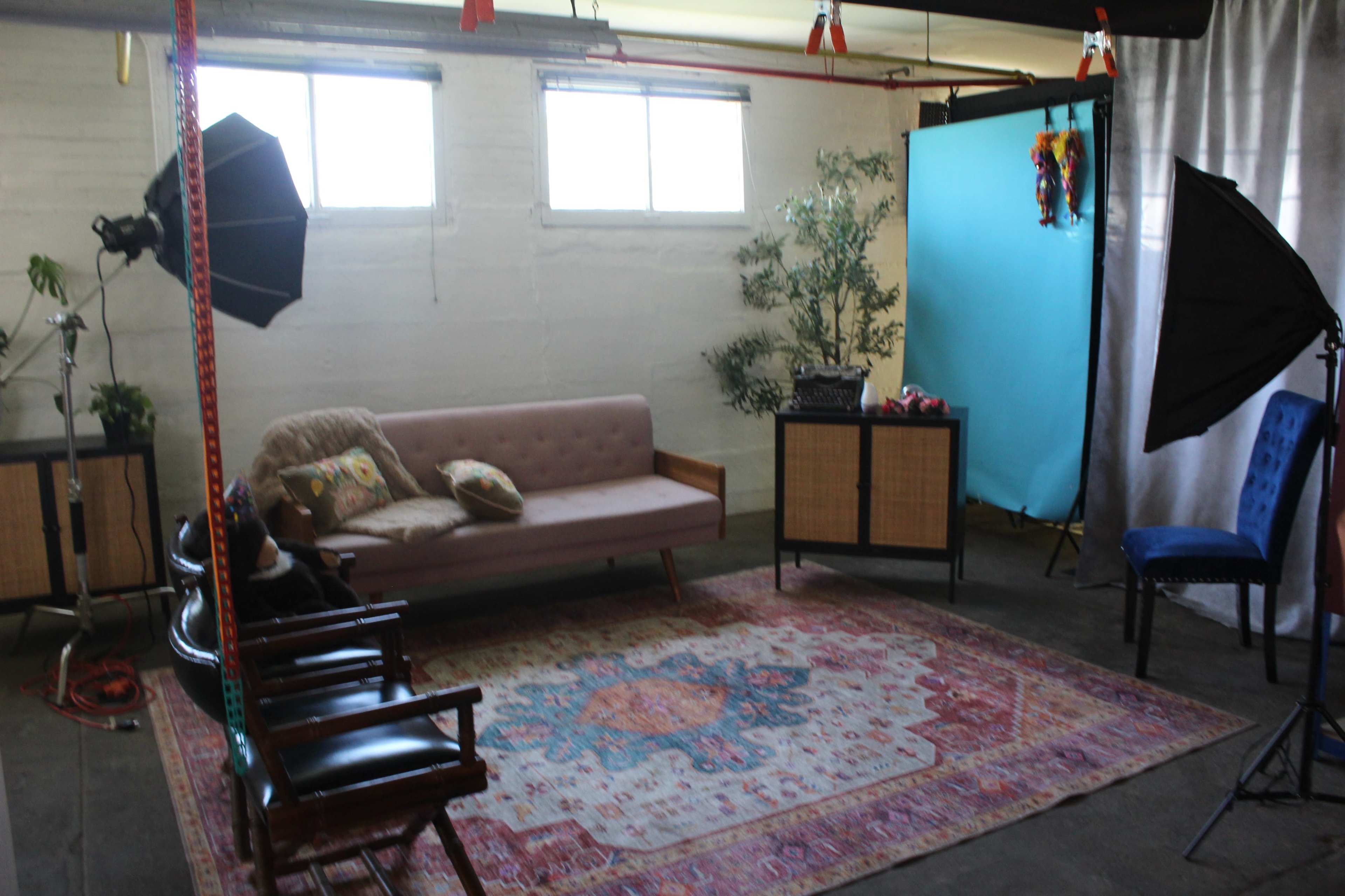 The image shows a photography studio featuring a pink sofa, a rug, two chairs, and a blue backdrop, with various lighting equipment and decorative plants.