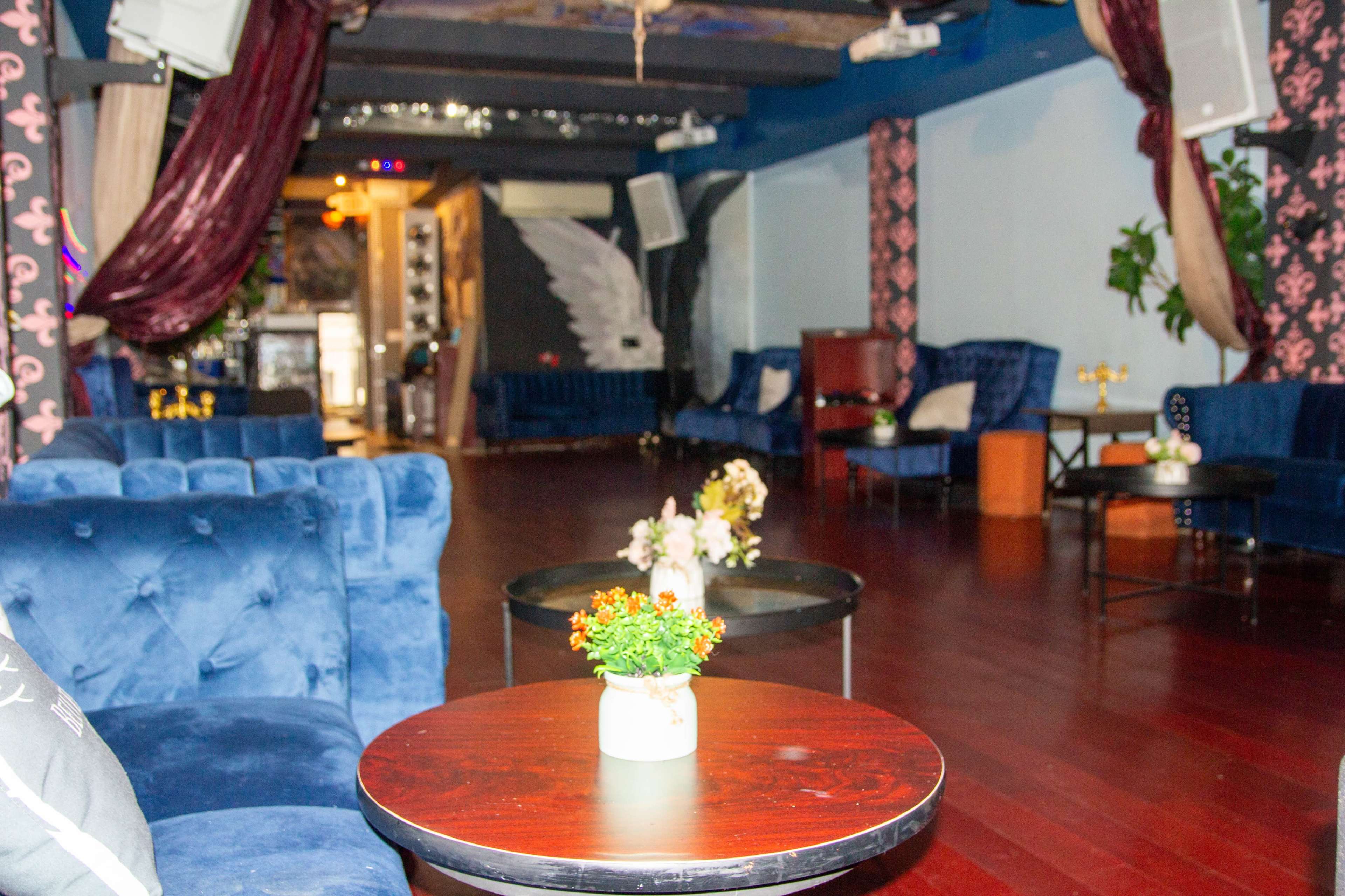 The image shows a sparsely furnished lounge area with blue sofas, small tables featuring potted flowers, and a polished wooden floor.