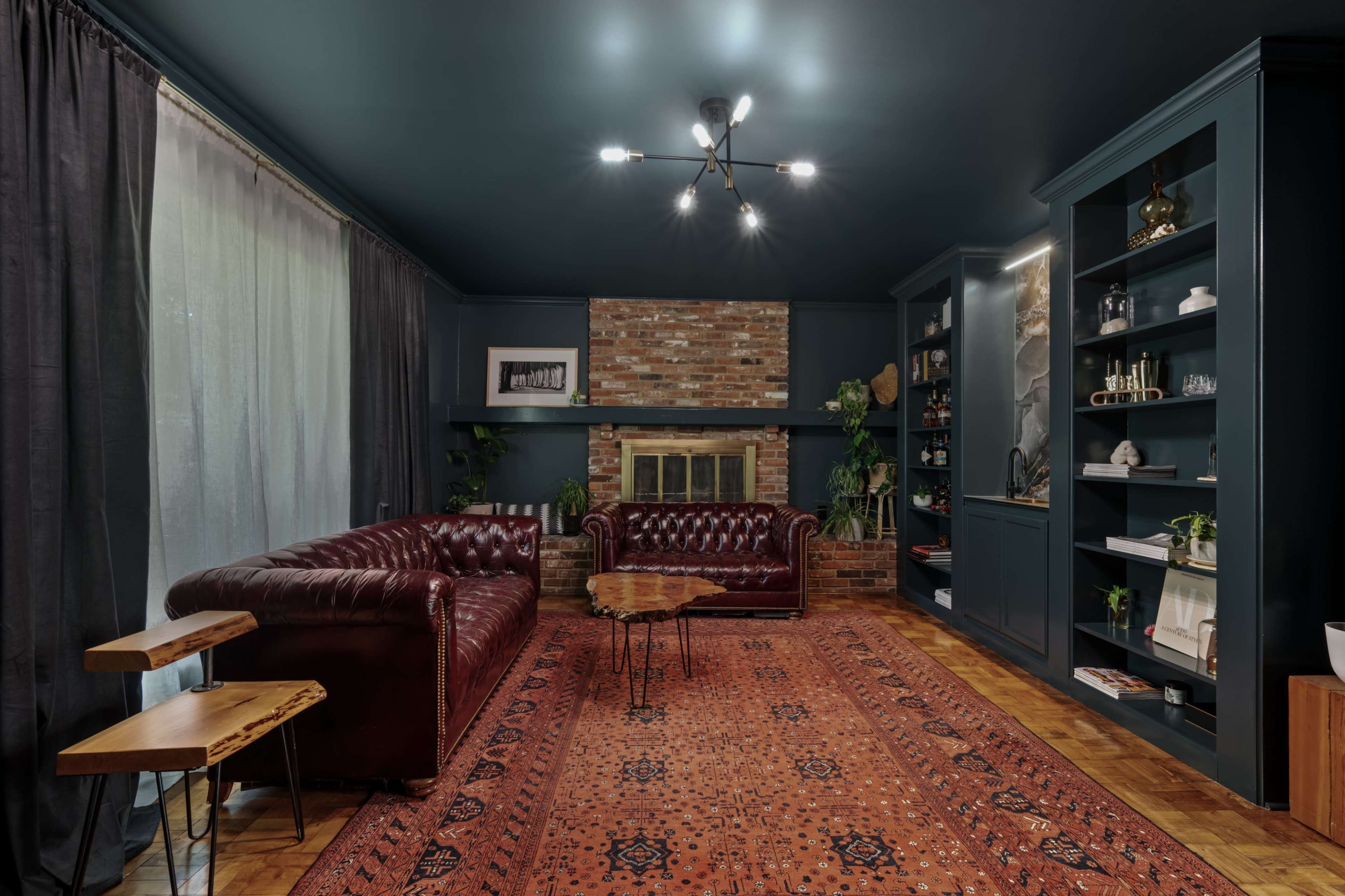 A cozy living room featuring a dark blue ceiling, a brick fireplace, a burgundy leather sofa, and a patterned area rug.