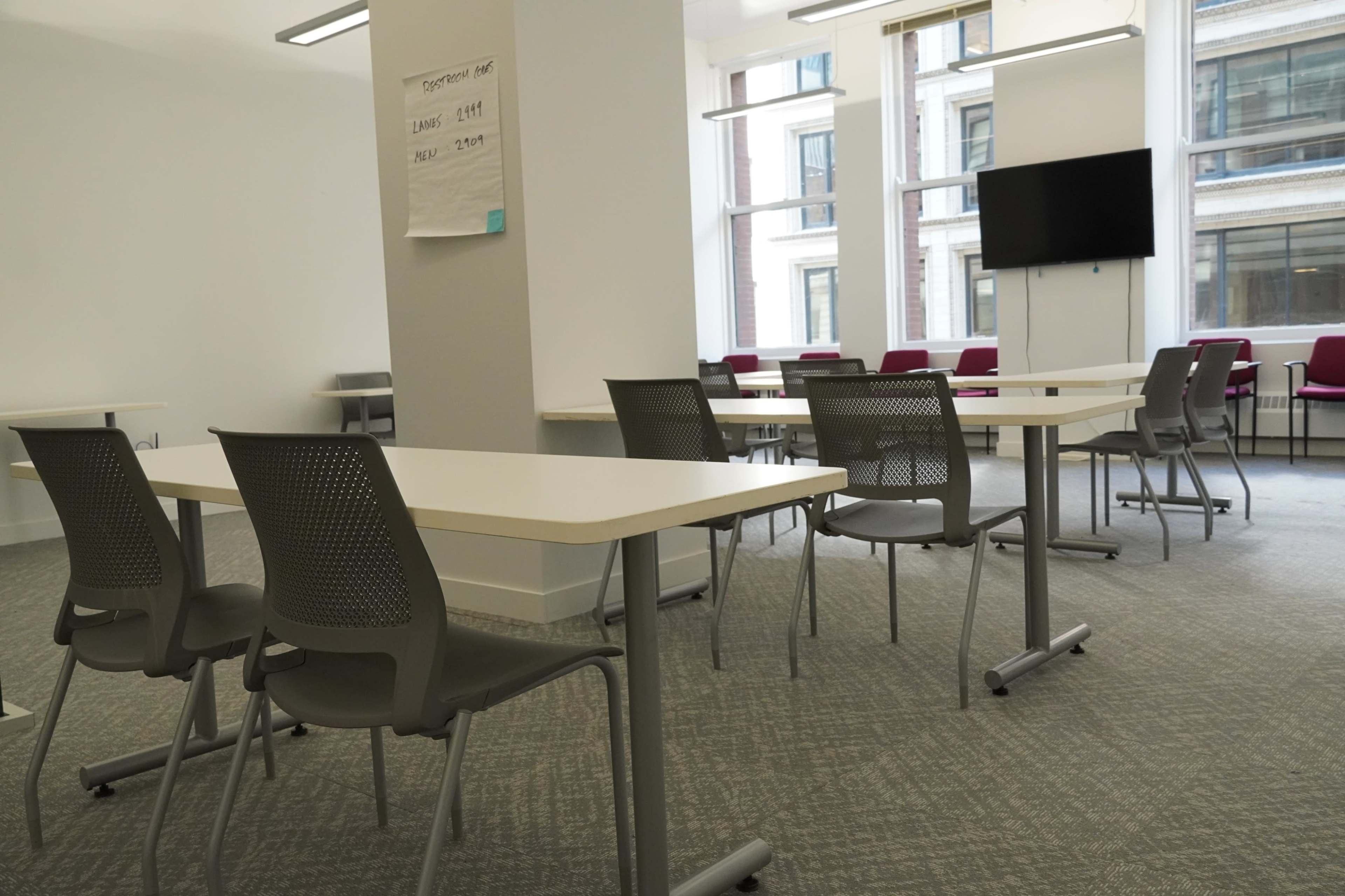 A modern, empty classroom with several tables and chairs arranged around the room, featuring large windows and a wall-mounted screen.