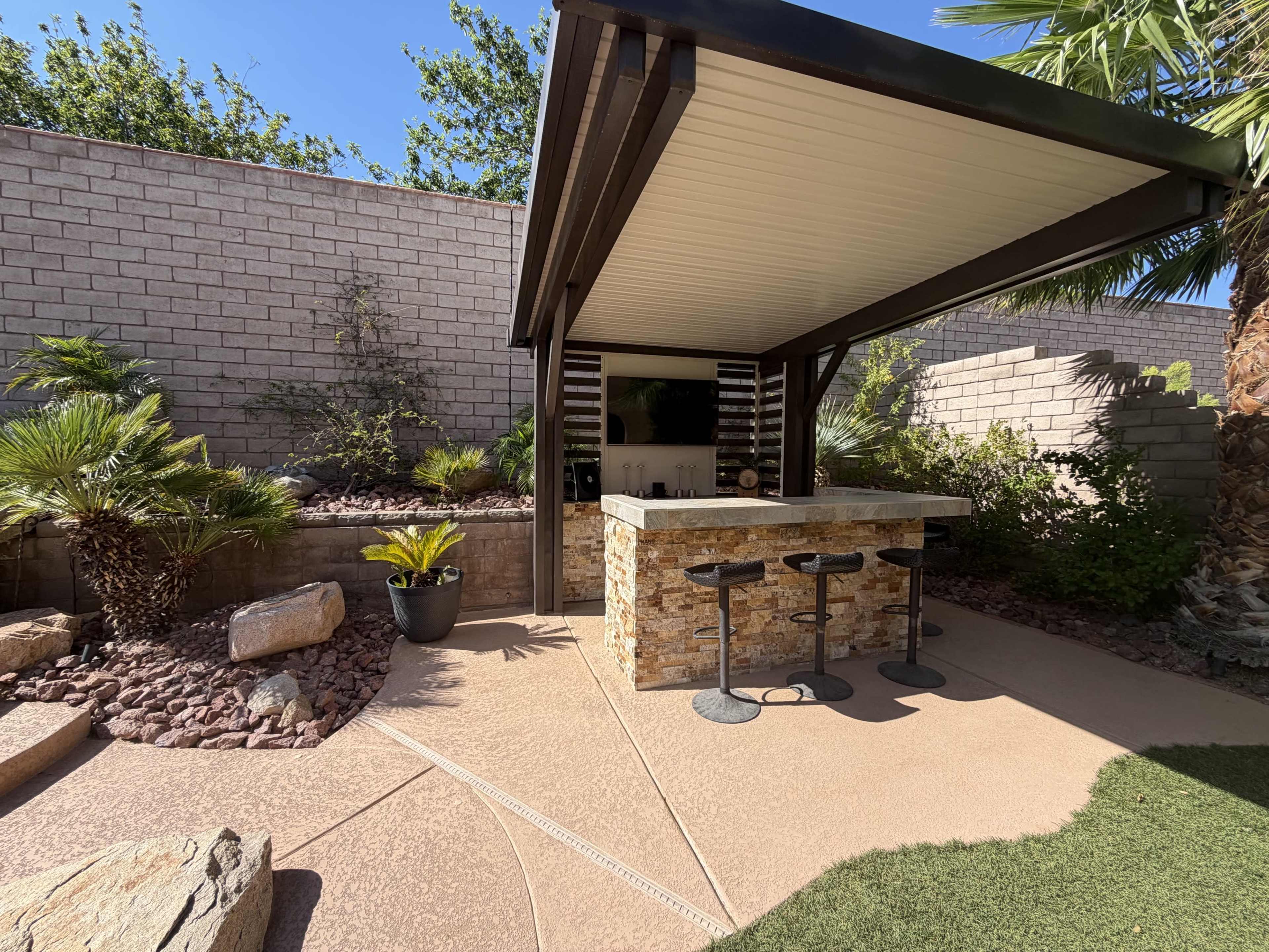 The image shows an outdoor bar area with a stone countertop and metal stools, surrounded by landscaped rocks and plants under a shaded structure.