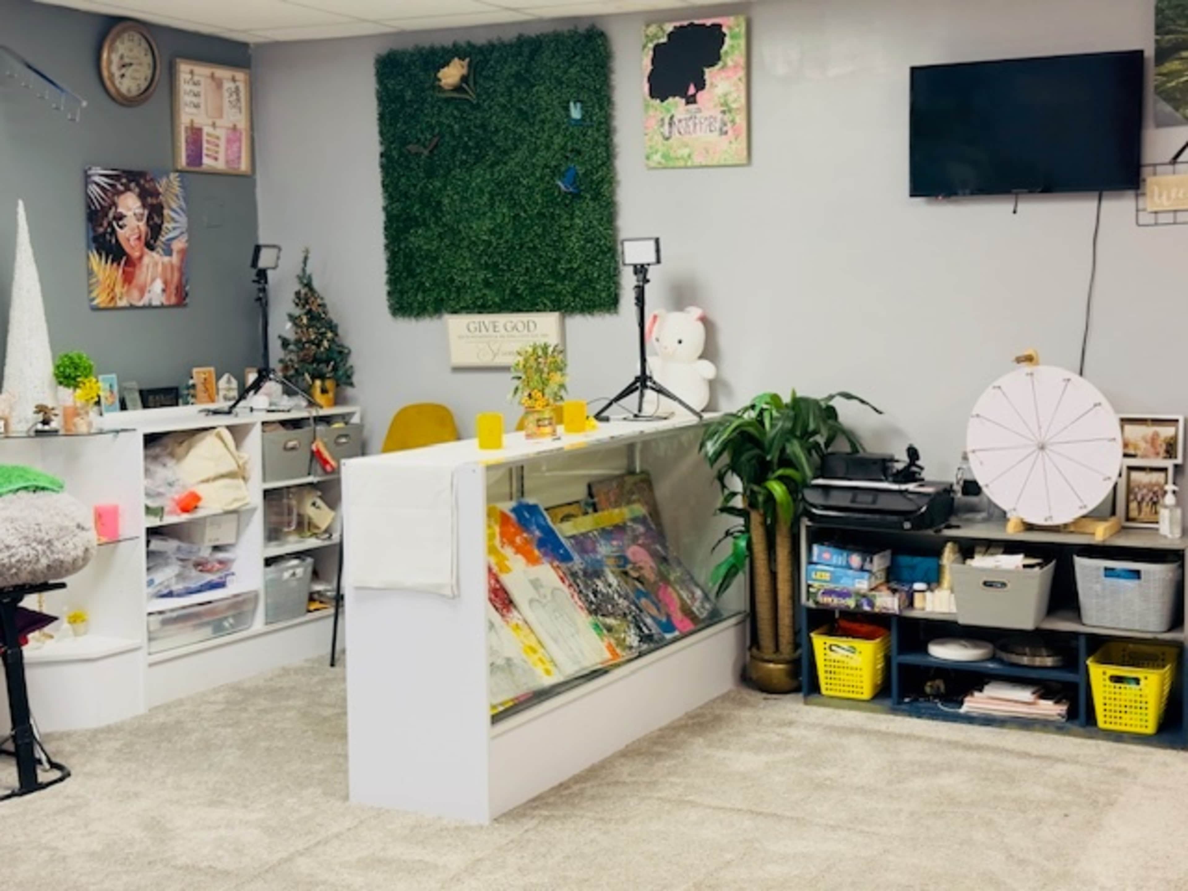 The image shows an indoor creative workspace featuring a display counter with art supplies, a green wall decoration, and various organizational bins.