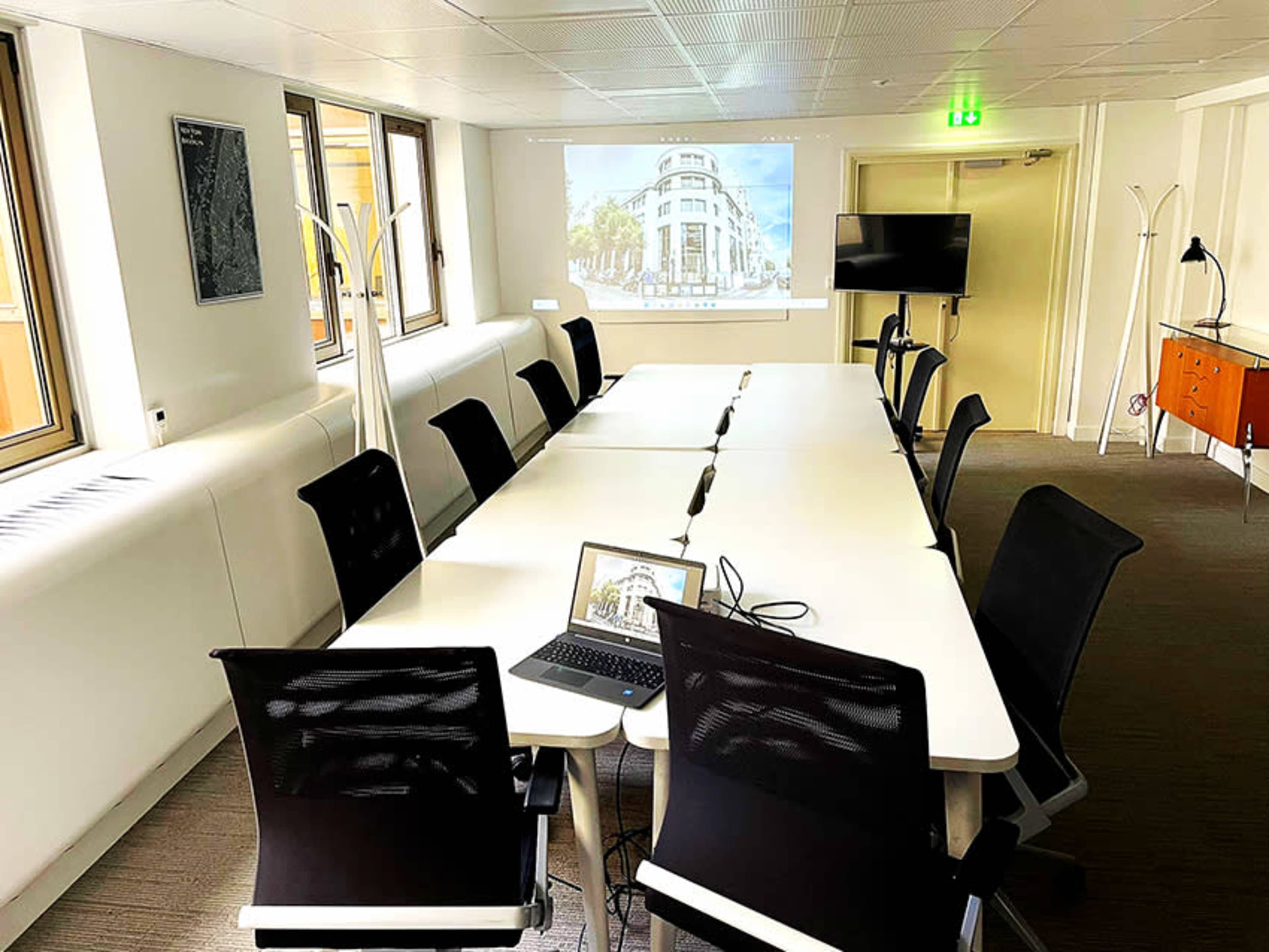 A modern conference room features a long table surrounded by black chairs, with a laptop on the table and a projector displays an image on the wall.