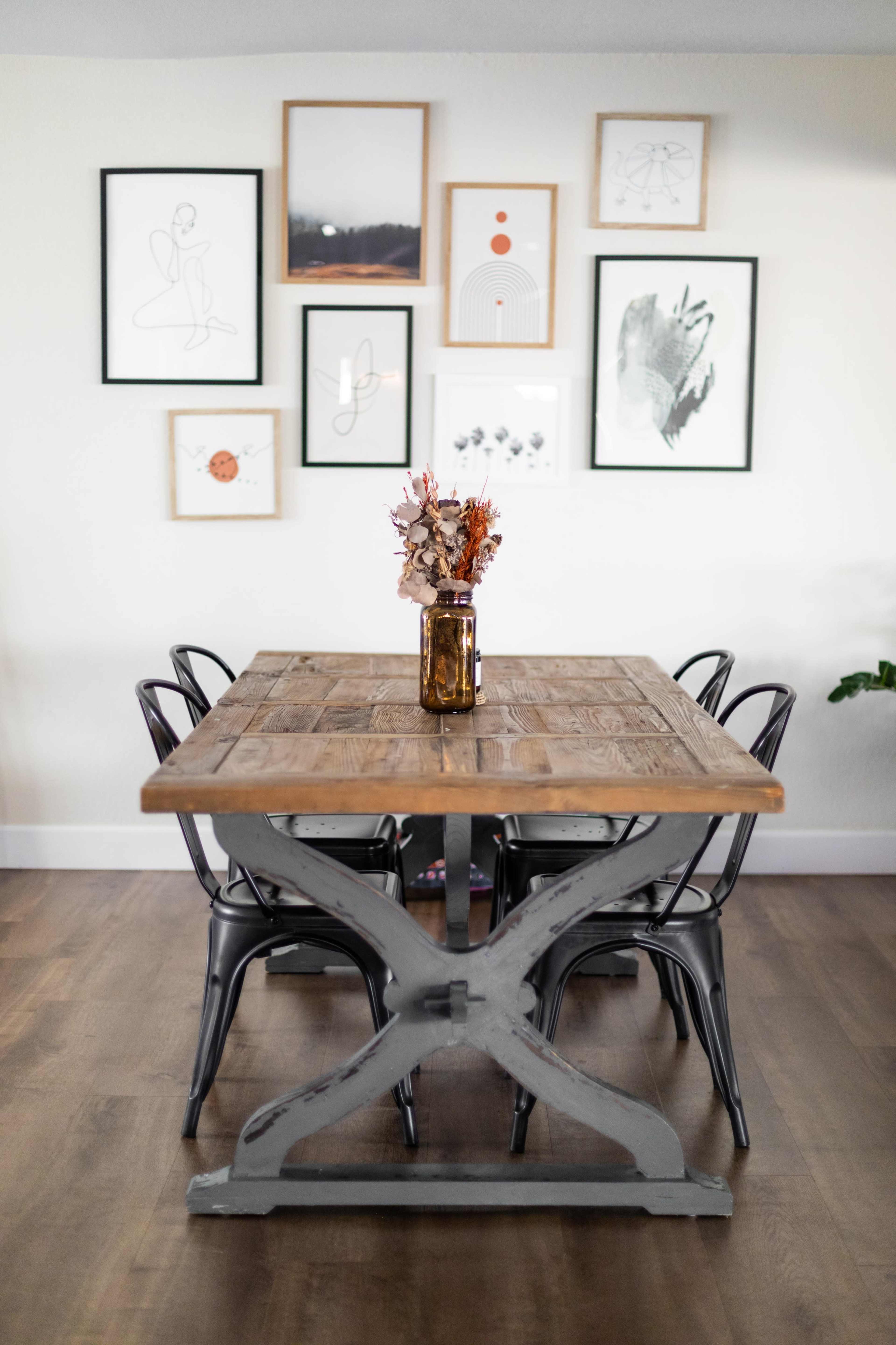 A wooden dining table with a metal base is surrounded by four chairs and positioned in front of a wall decorated with framed artwork.
