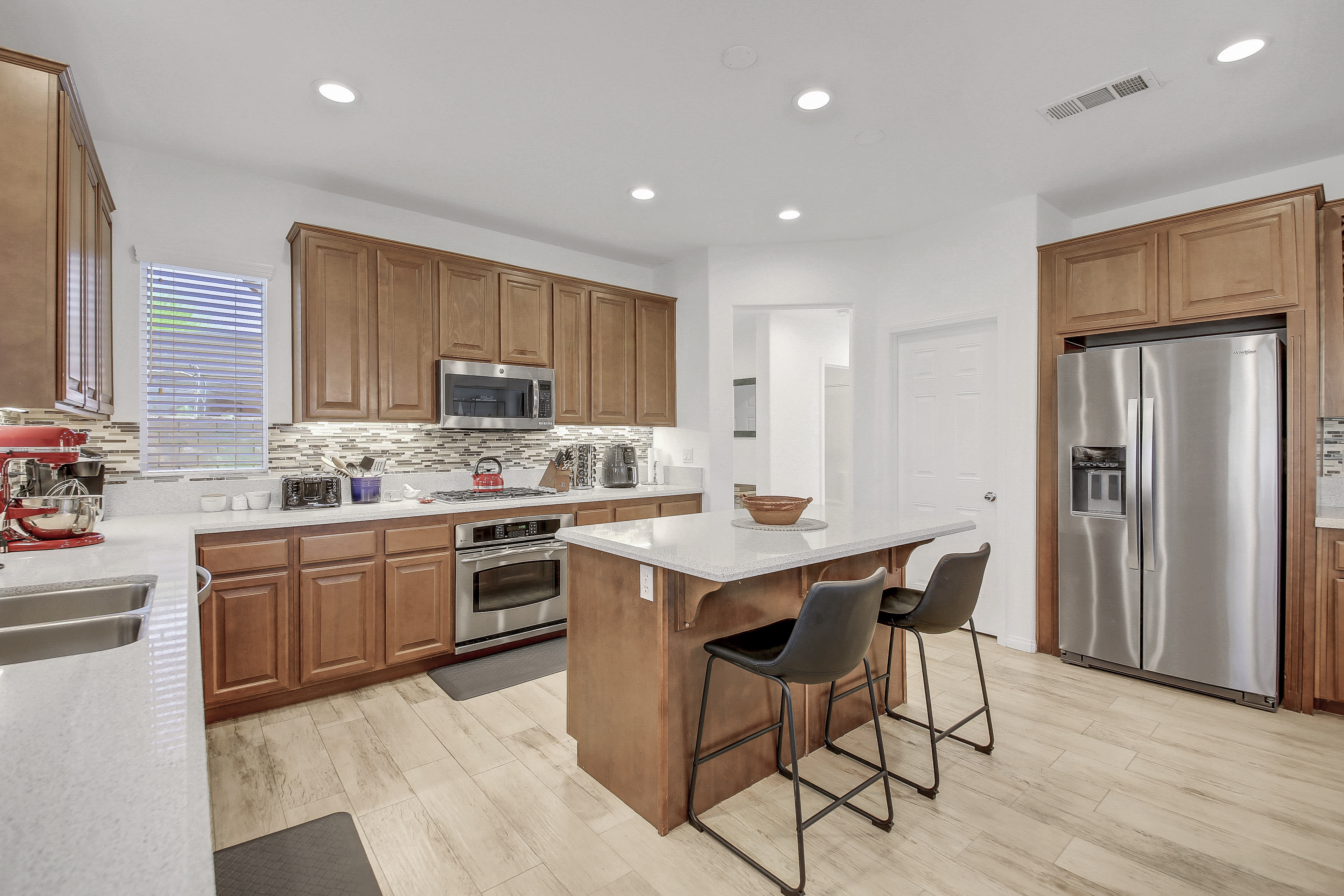 The image shows a modern kitchen featuring wooden cabinets, stainless steel appliances, and an island with seating for three.