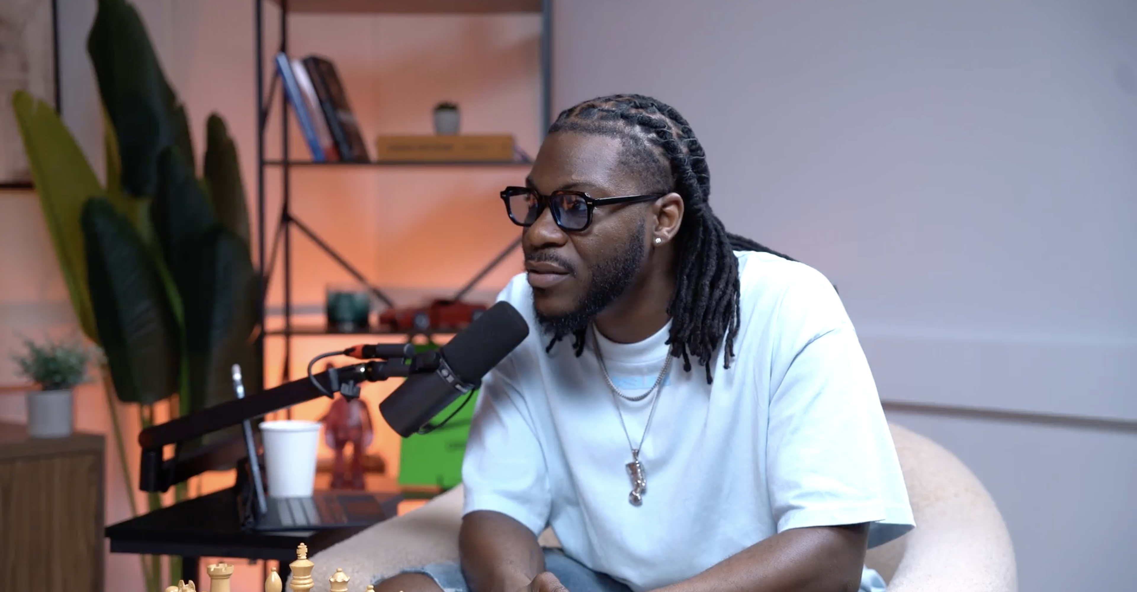 A man with dreadlocks is sitting near a microphone and a chess set, facing the camera in a modern, well-lit room.