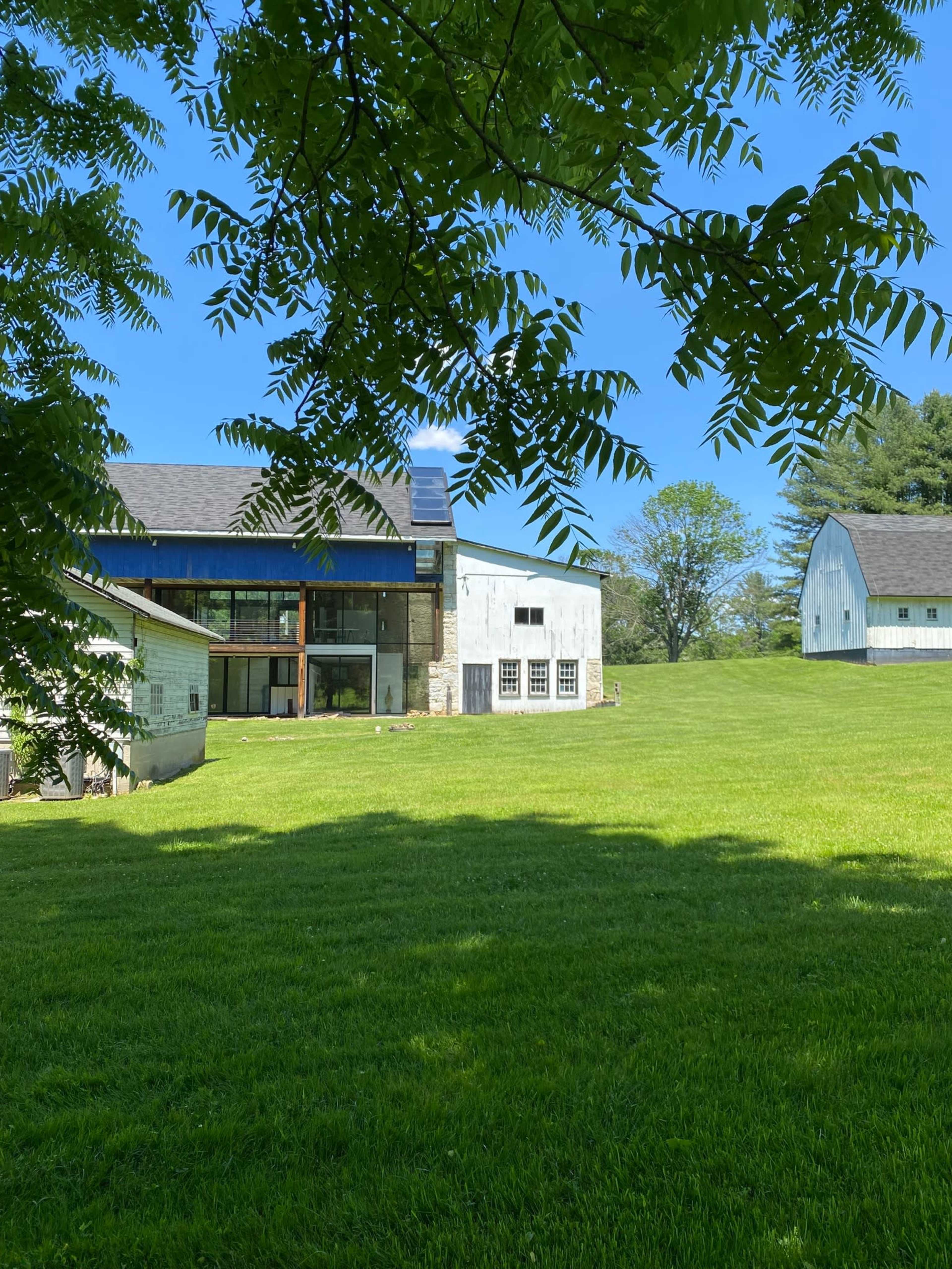A modern house with large windows stands next to two barns on a landscaped green lawn under a clear blue sky.