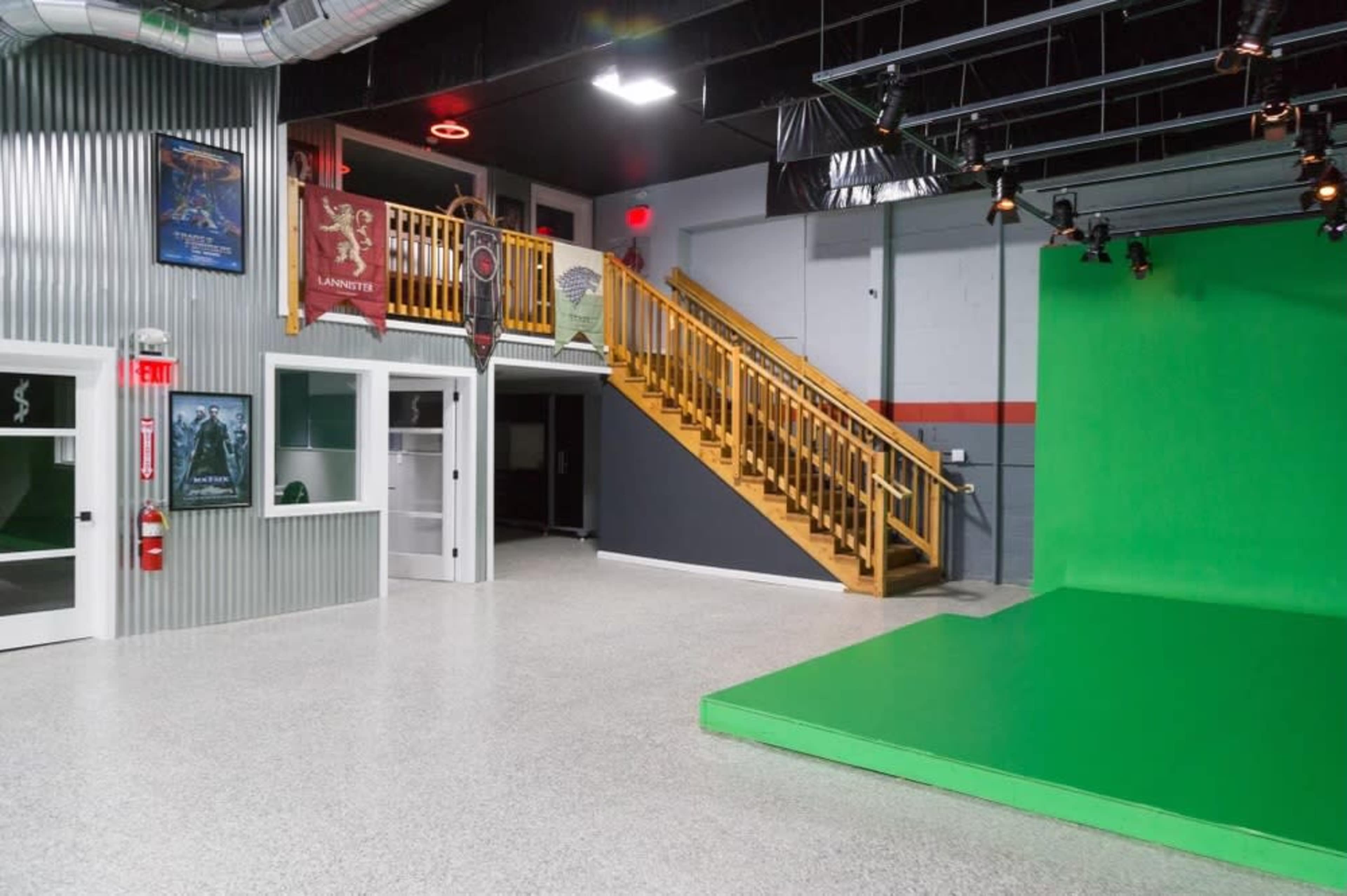A spacious film studio with a green screen area, a staircase leading to an upper level, and various posters on the walls.