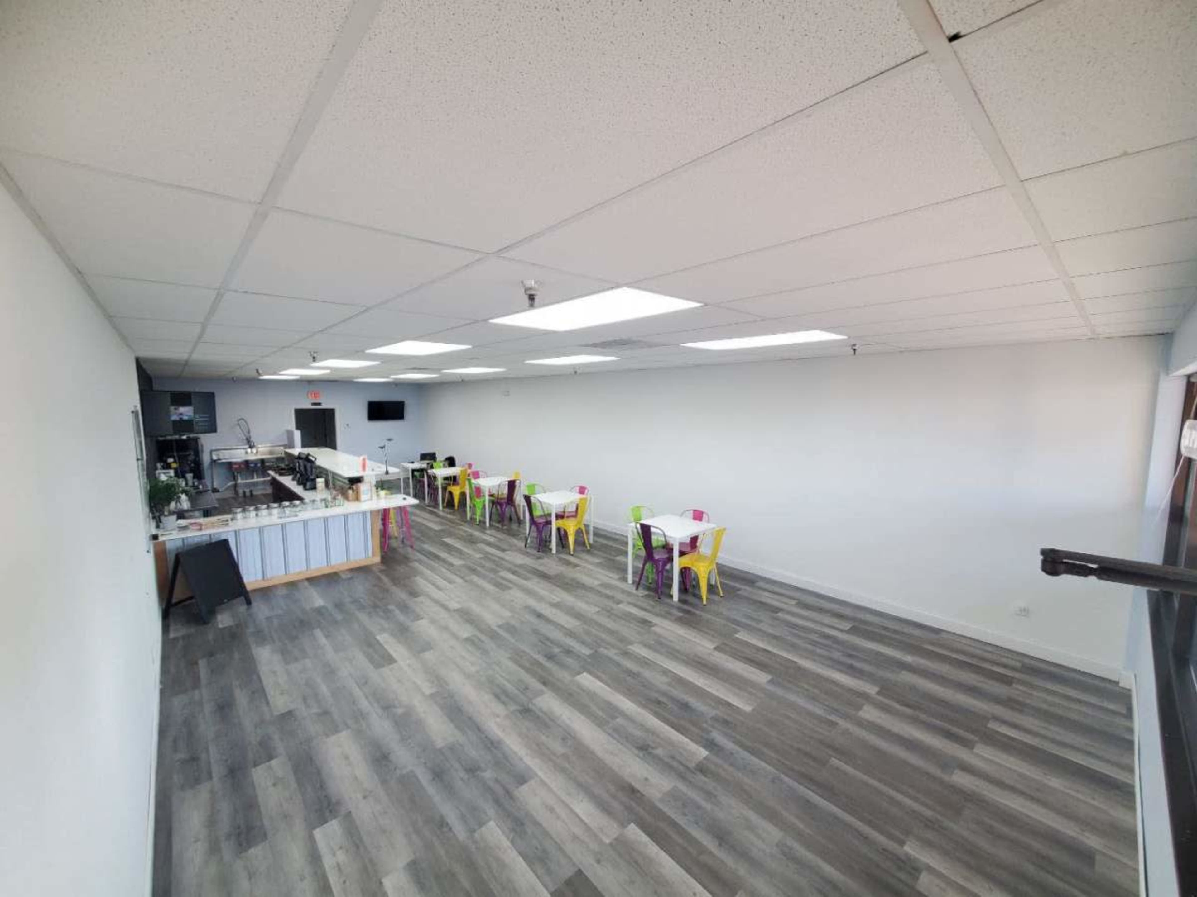 A spacious, minimally furnished interior of a dining area with a row of colorful chairs and tables arranged neatly on the gray floor.