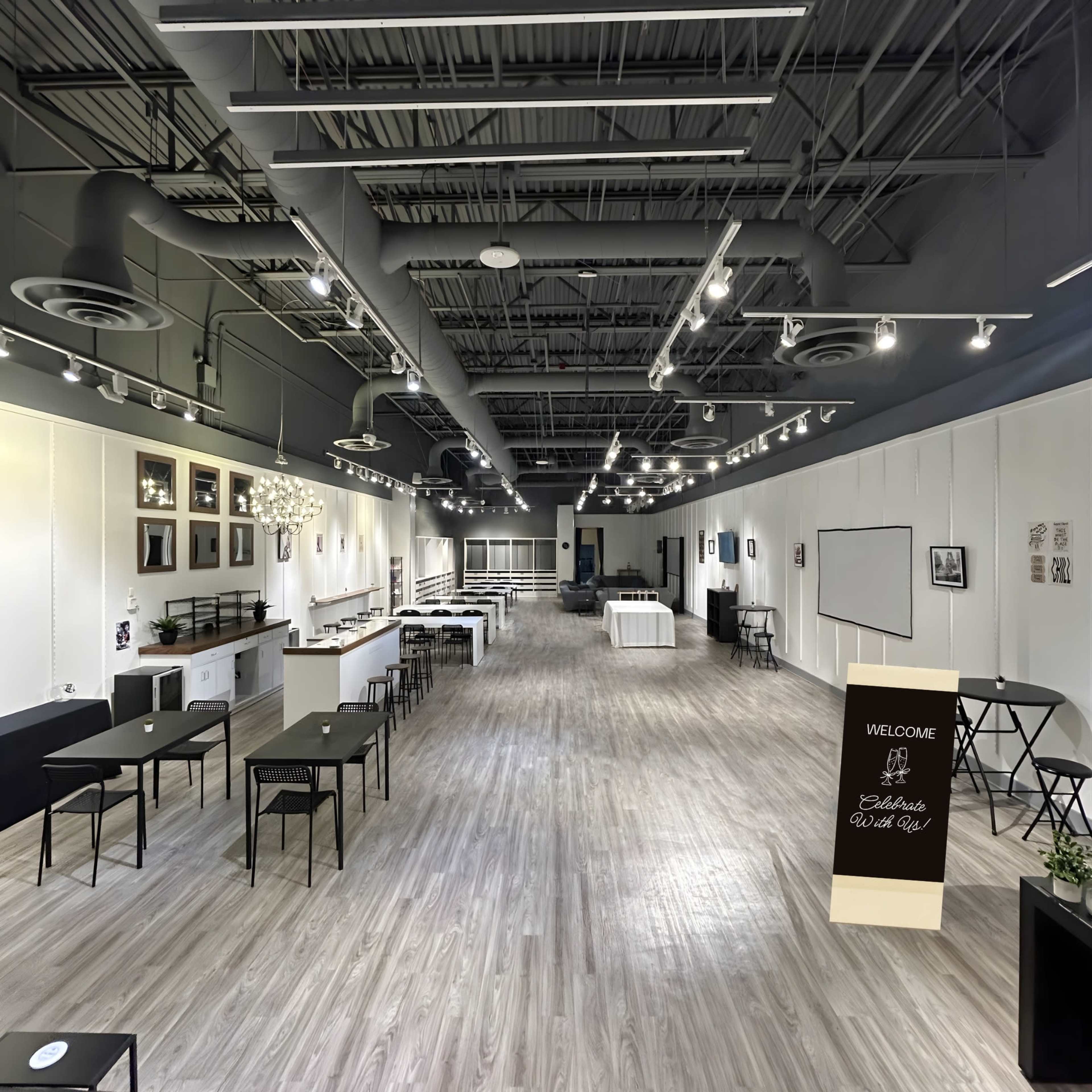 The image shows a spacious indoor venue with light-colored wooden flooring, featuring several rectangular tables and chairs arranged neatly, along with wall-mounted decorations and overhead lighting.