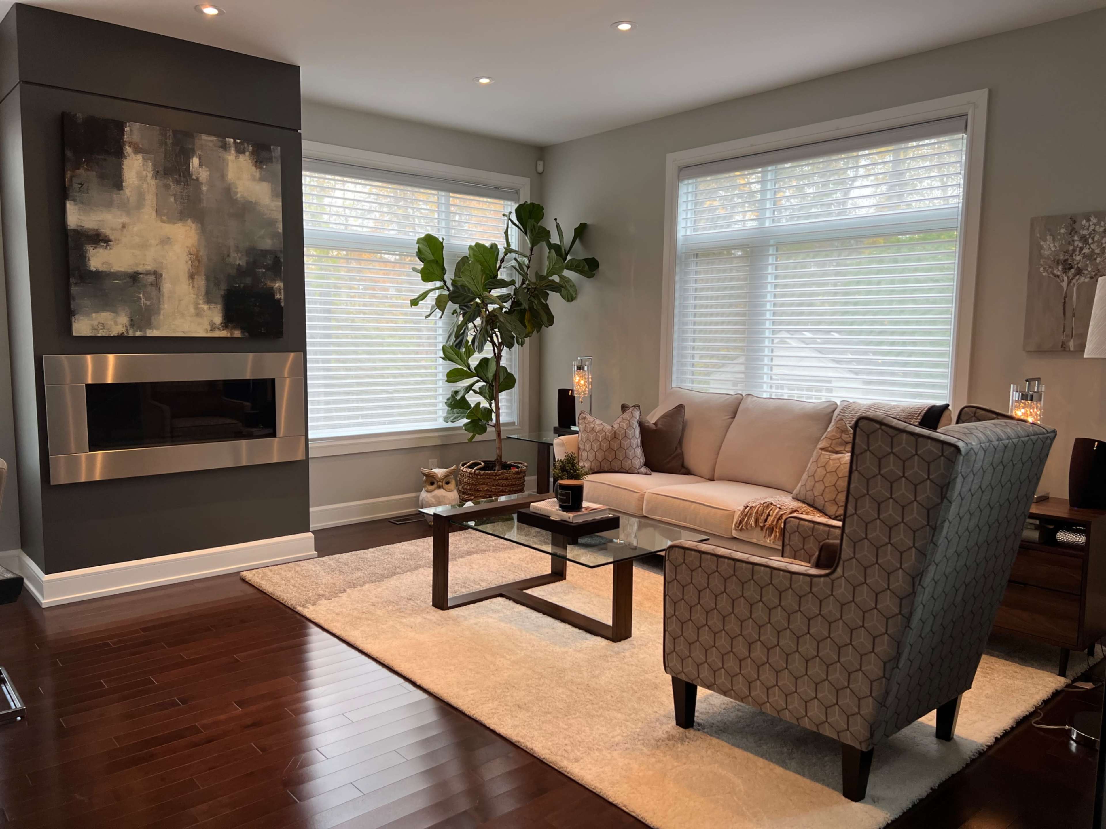 A modern living room with a sofa, coffee table, and large windows allowing natural light, complemented by a decorative plant and a wall-mounted fireplace.