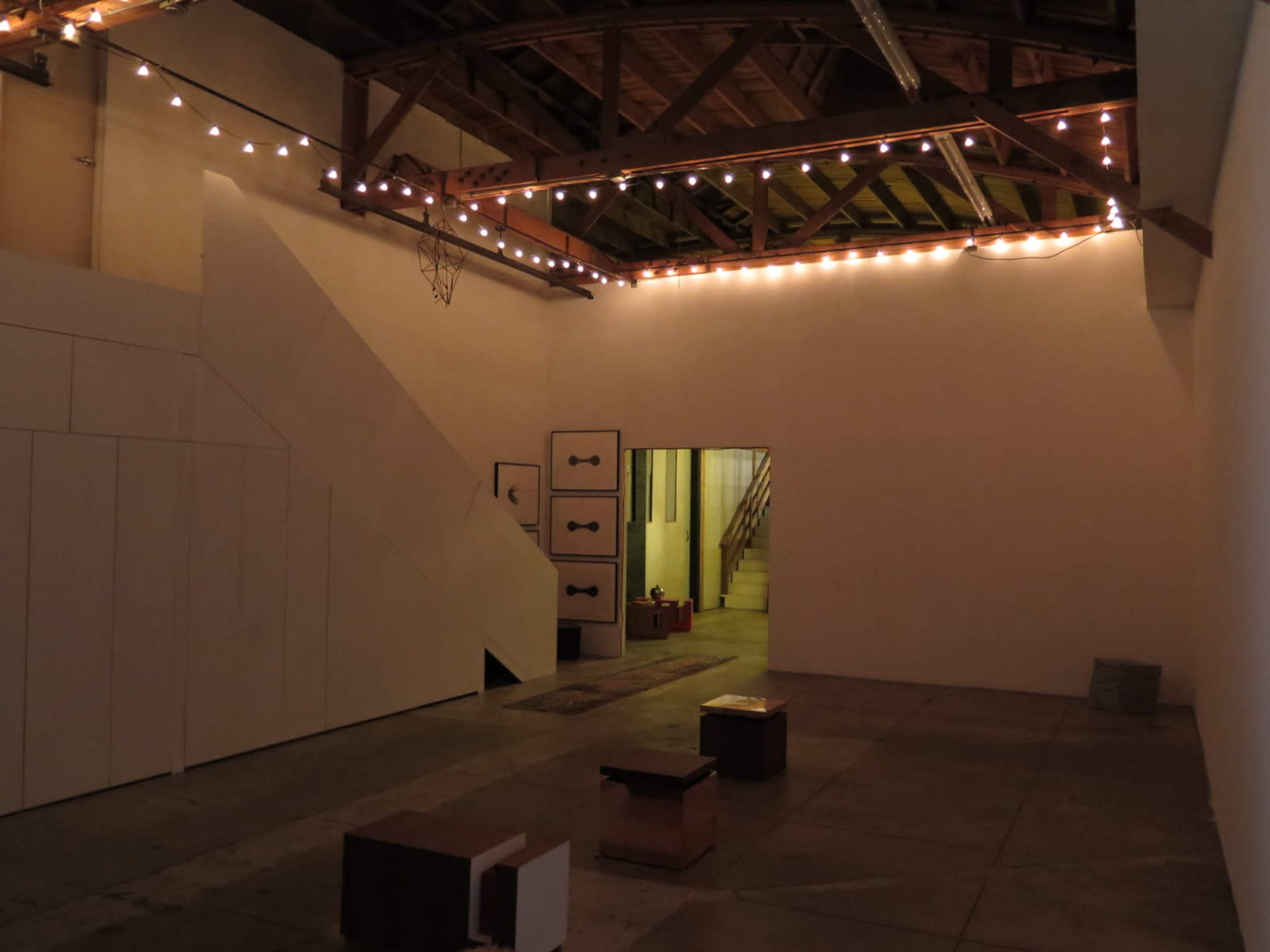 An empty warehouse space with exposed beams, string lights along the ceiling, and a staircase leading to an upper level.