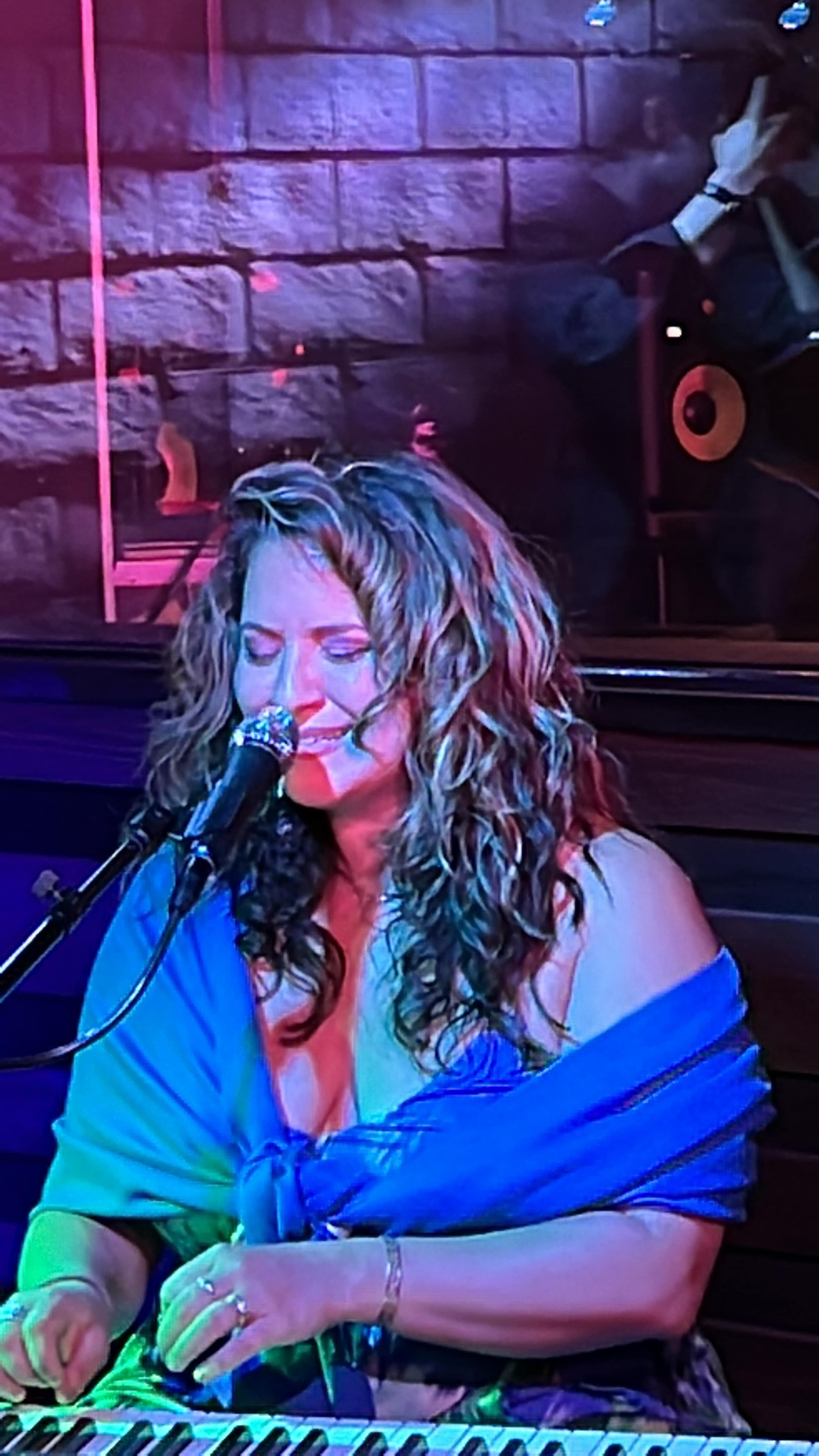 A woman with curly hair plays a keyboard while singing into a microphone, illuminated by colorful stage lights.
