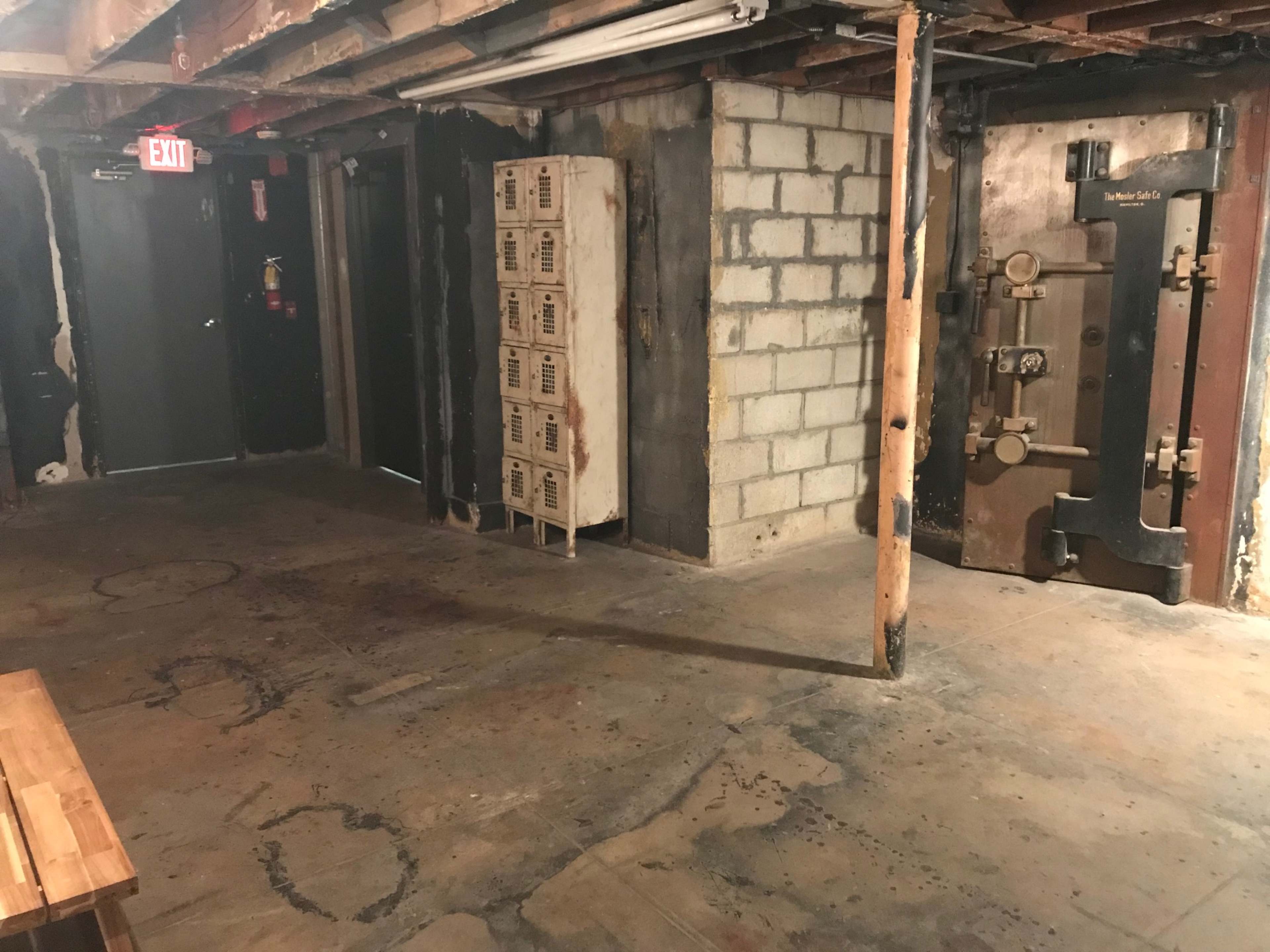 A dimly lit basement area with exposed brick, a large metal safe door, a set of lockers, and a concrete floor.