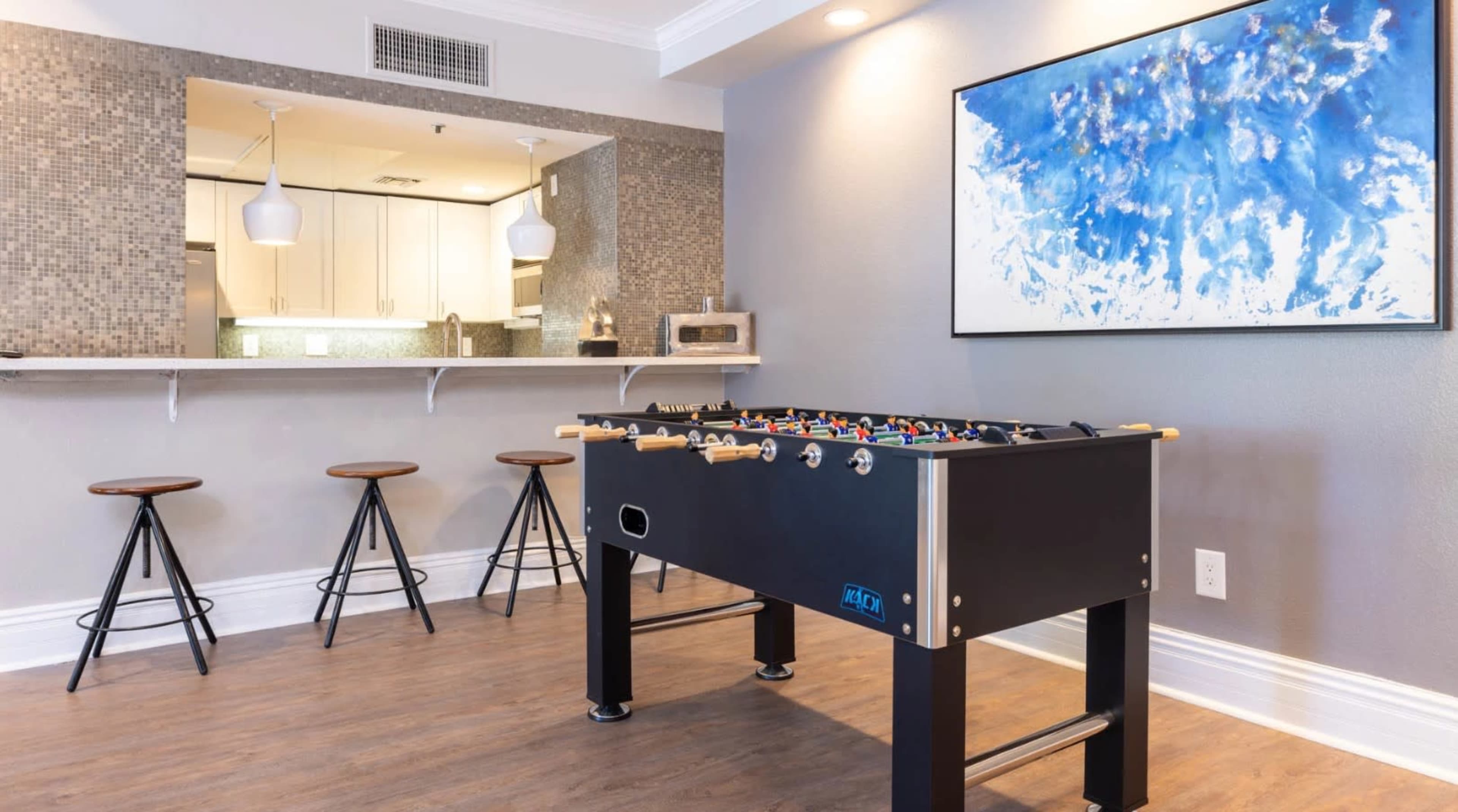 A modern game room features a foosball table in the foreground, with a bar area and stylish stools in the background.