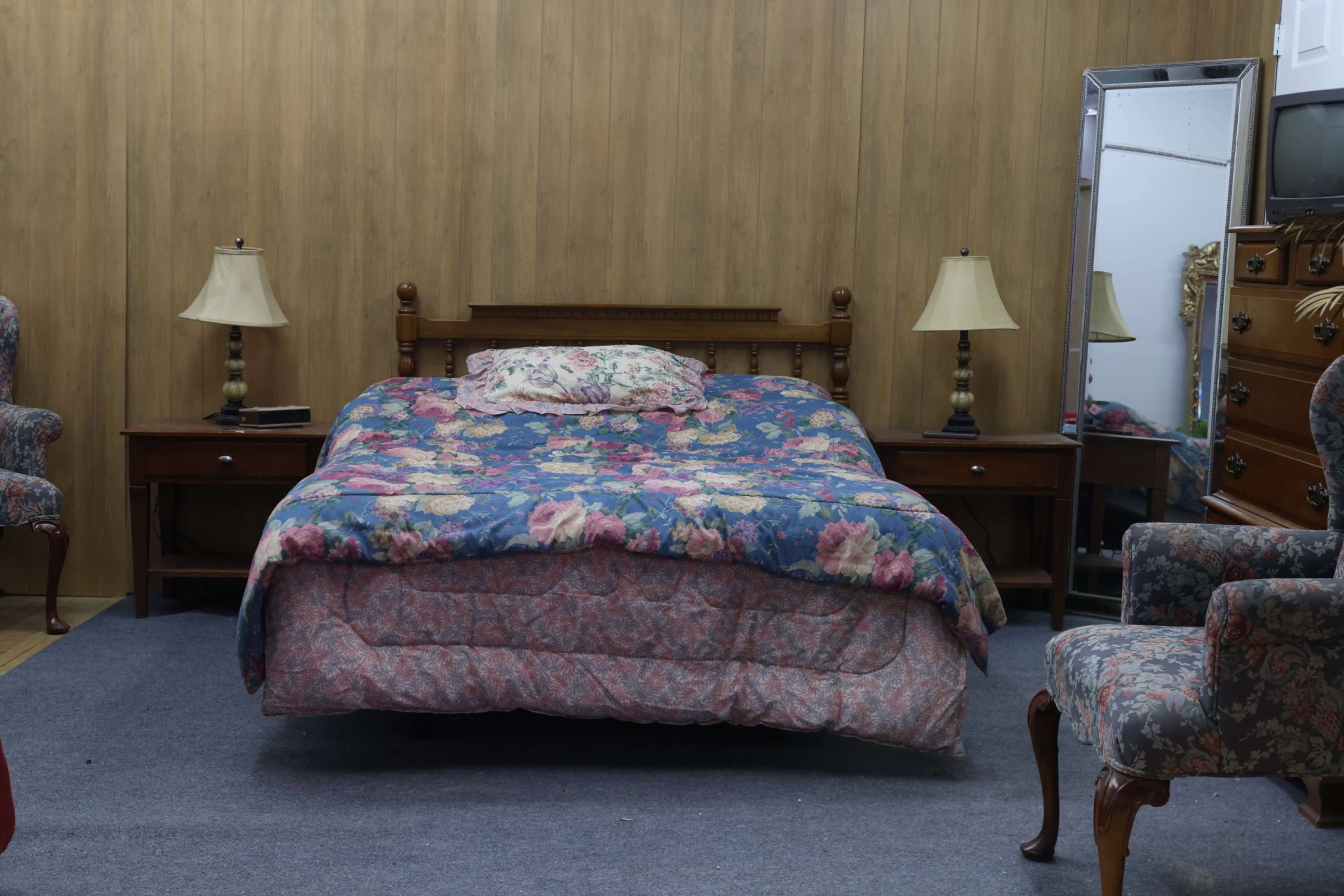The image shows a simple bedroom with a bed covered in a floral quilt, two matching bedside lamps, a mirror, an armchair, and a dresser against a wooden paneled wall.