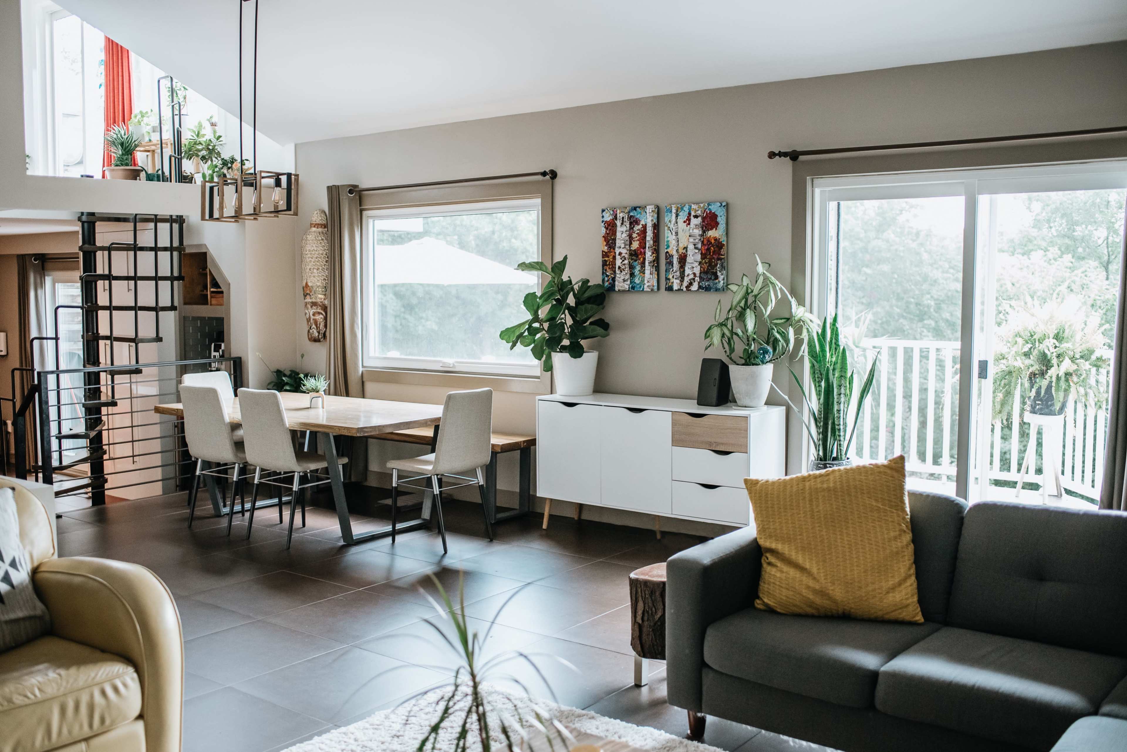 A modern living space featuring a dining area with a wooden table and chairs, a grey sofa, and large windows with plants on display.