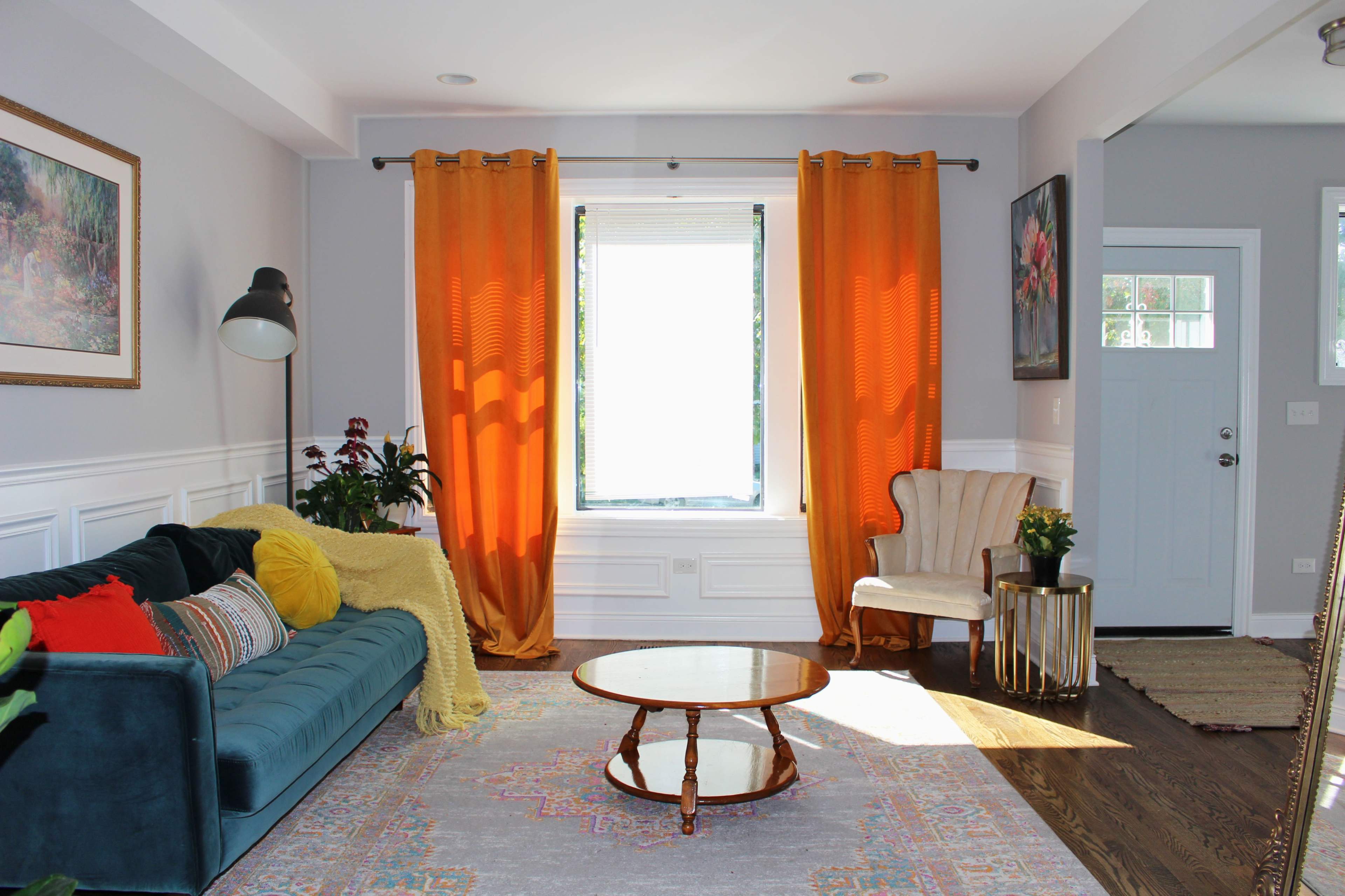 The image shows a brightly lit living room featuring a teal sofa, a round wooden coffee table, and a white armchair, with orange curtains framing a large window.