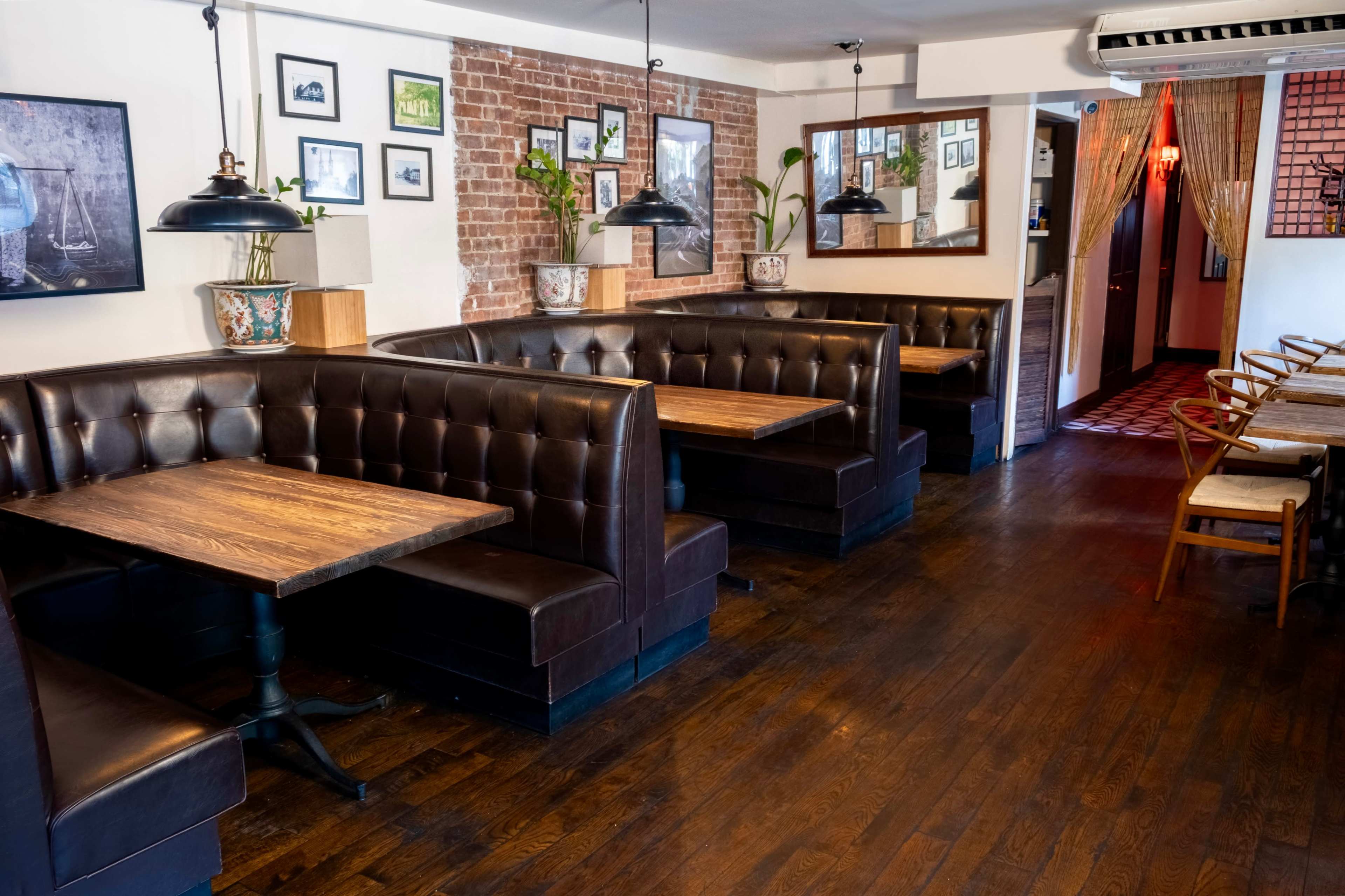 The image shows an empty restaurant interior with dark brown leather booths, wooden tables, and brick walls, adorned with framed pictures and plants.